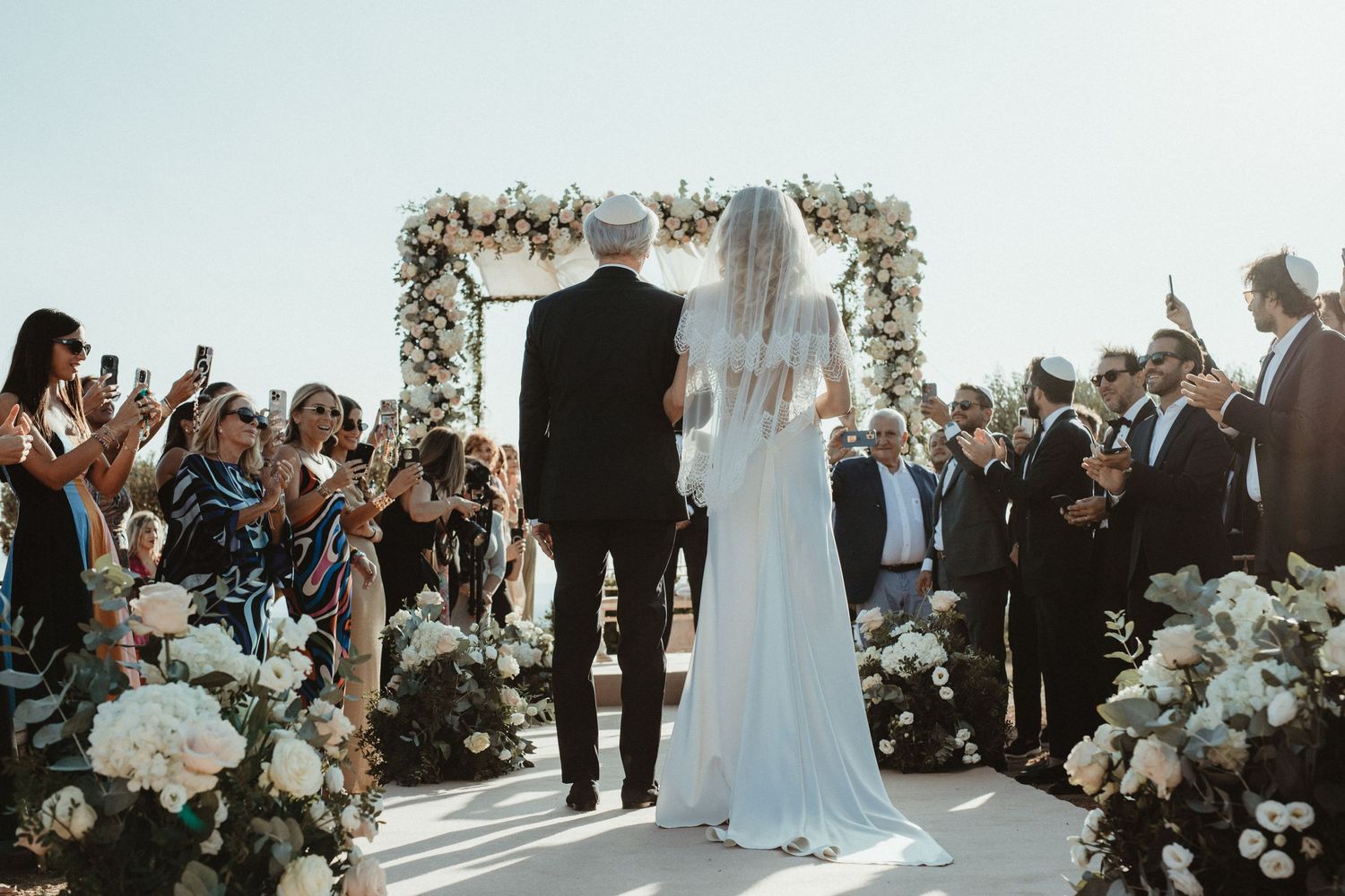 Floral arch decorated with white flowers stands against blue sky at outdoor ceremony.