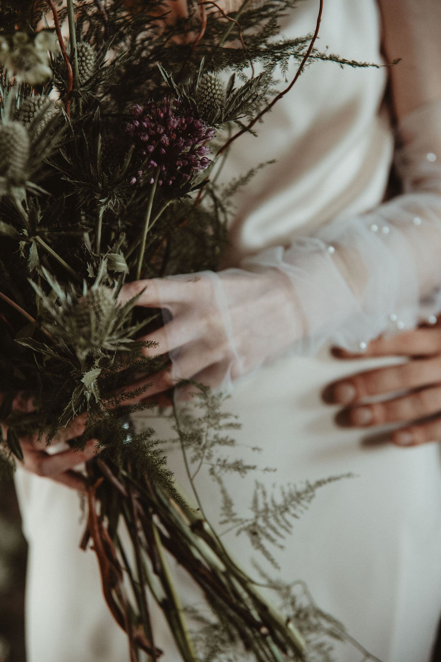 Close-up detail shot of hands holding a rustic bouquet with ferns and wildflowers.