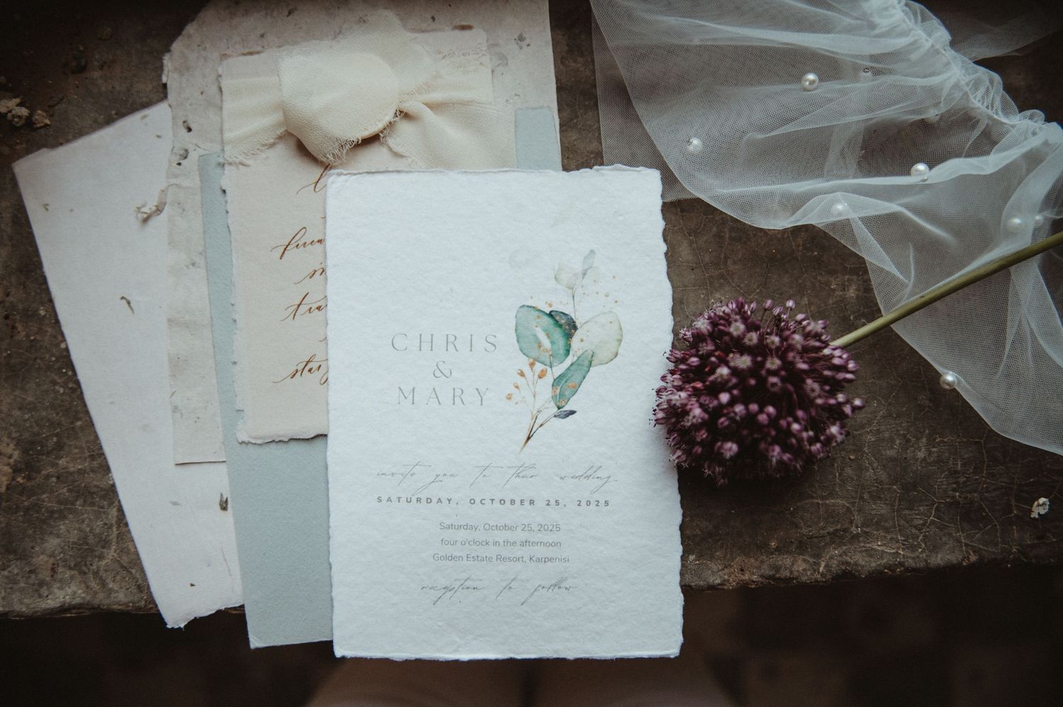Wedding stationery with delicate green leaf design and dried flowers on dark surface.