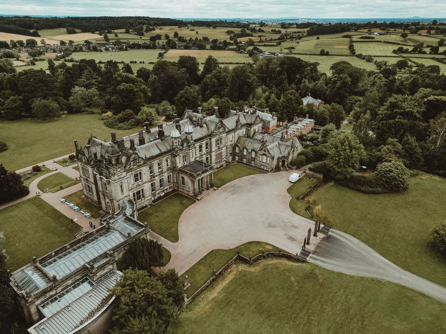 An aerial drone view of a grand manor estate with extensive gardens and winding driveways in the English countryside.