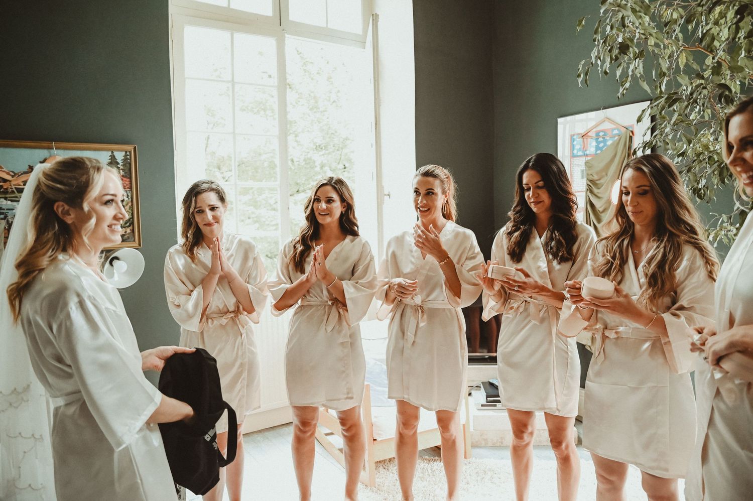Bridesmaids in white robes celebrate joyfully in bright, window-lit room with greenery.