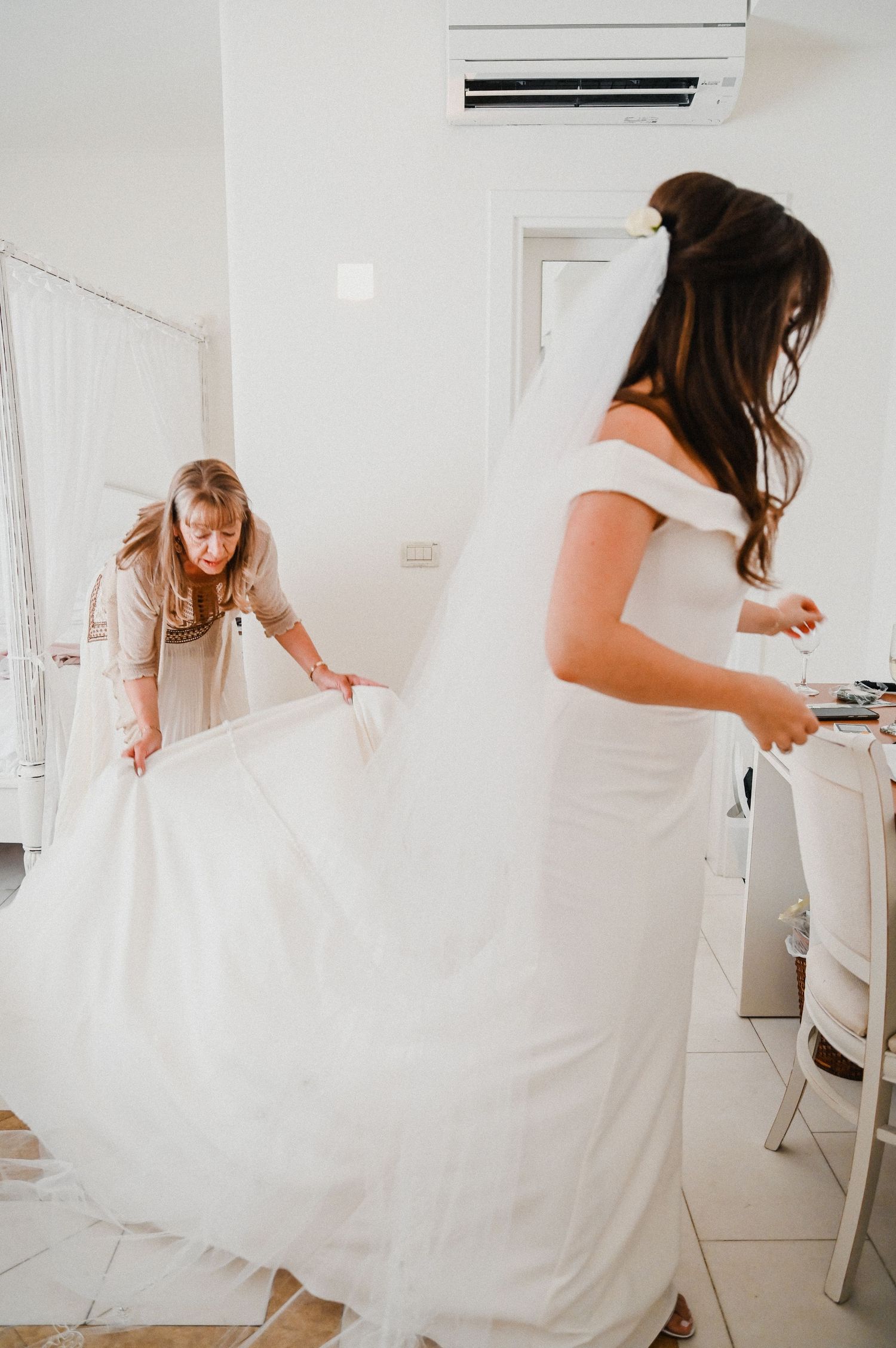 Two people preparing a flowing white wedding dress in a bright interior room.