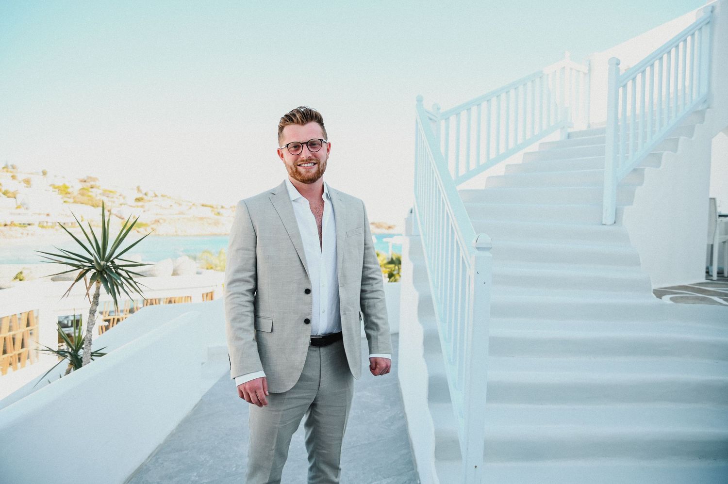 Groomsman in light gray suit stands on white stairs with scenic coastal view.