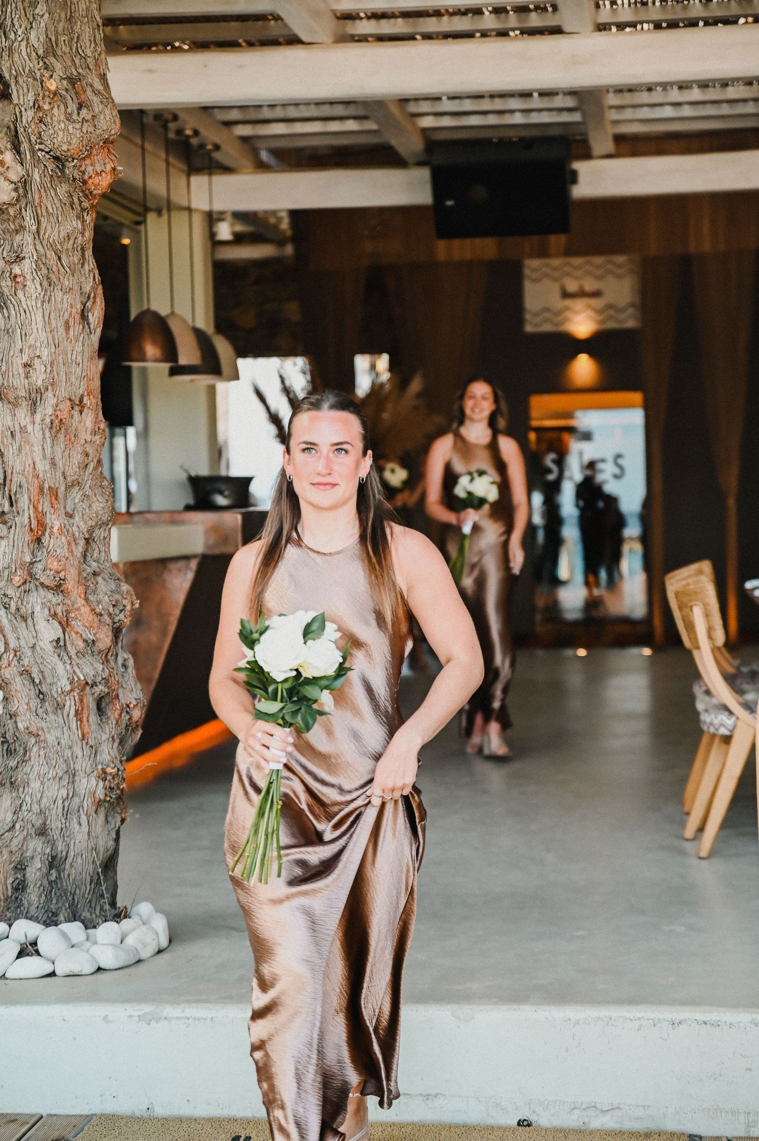 Wedding attendant in champagne silk dress holds white bouquet in modern indoor venue.
