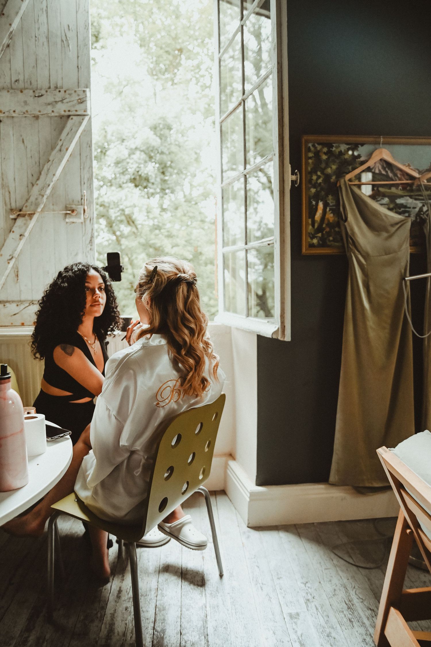 Getting ready scene by a bright window with a formal dress hanging in the background.