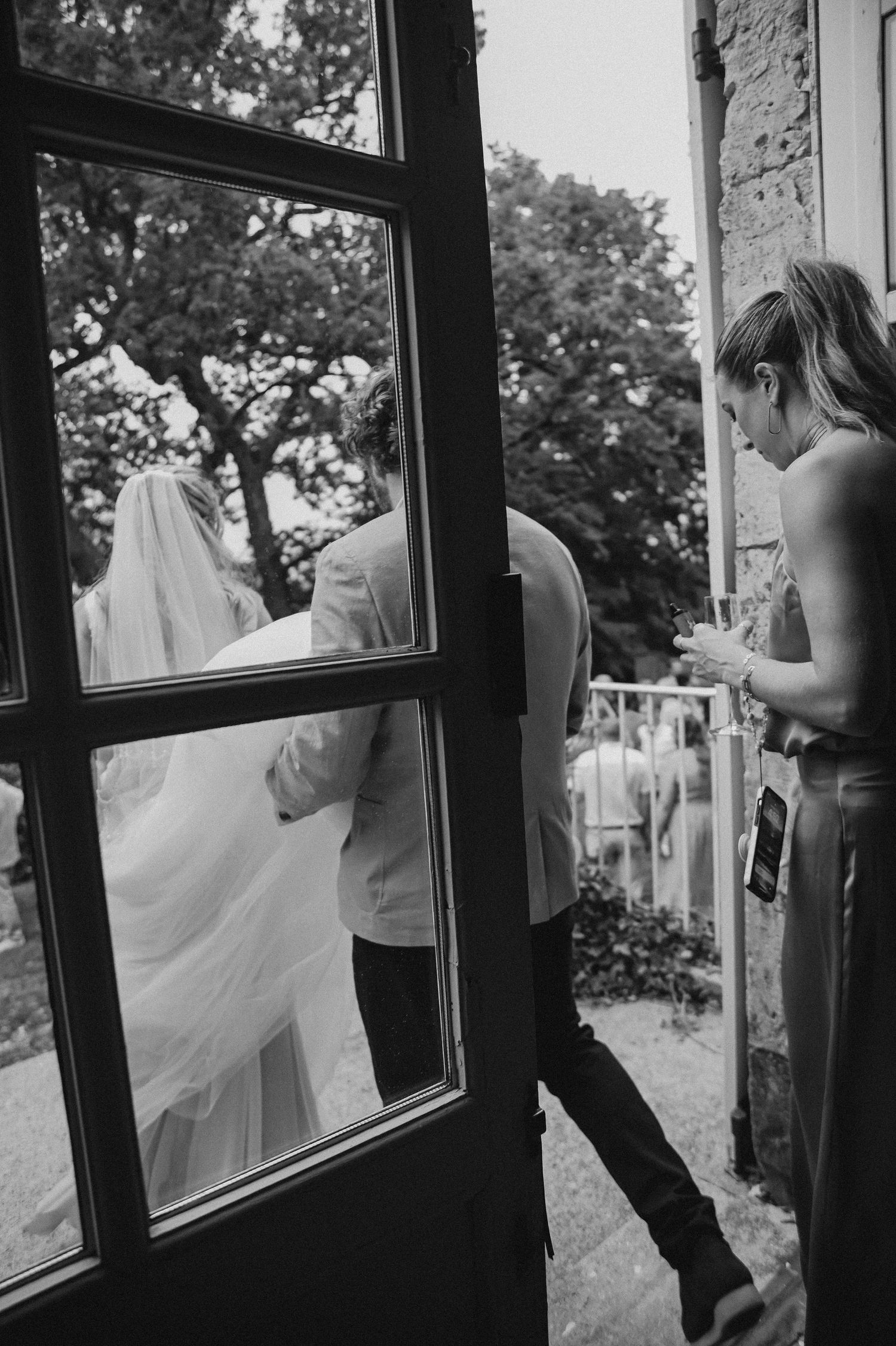 A black and white wedding photo showing a person through a window pane with flowing white fabric visible.