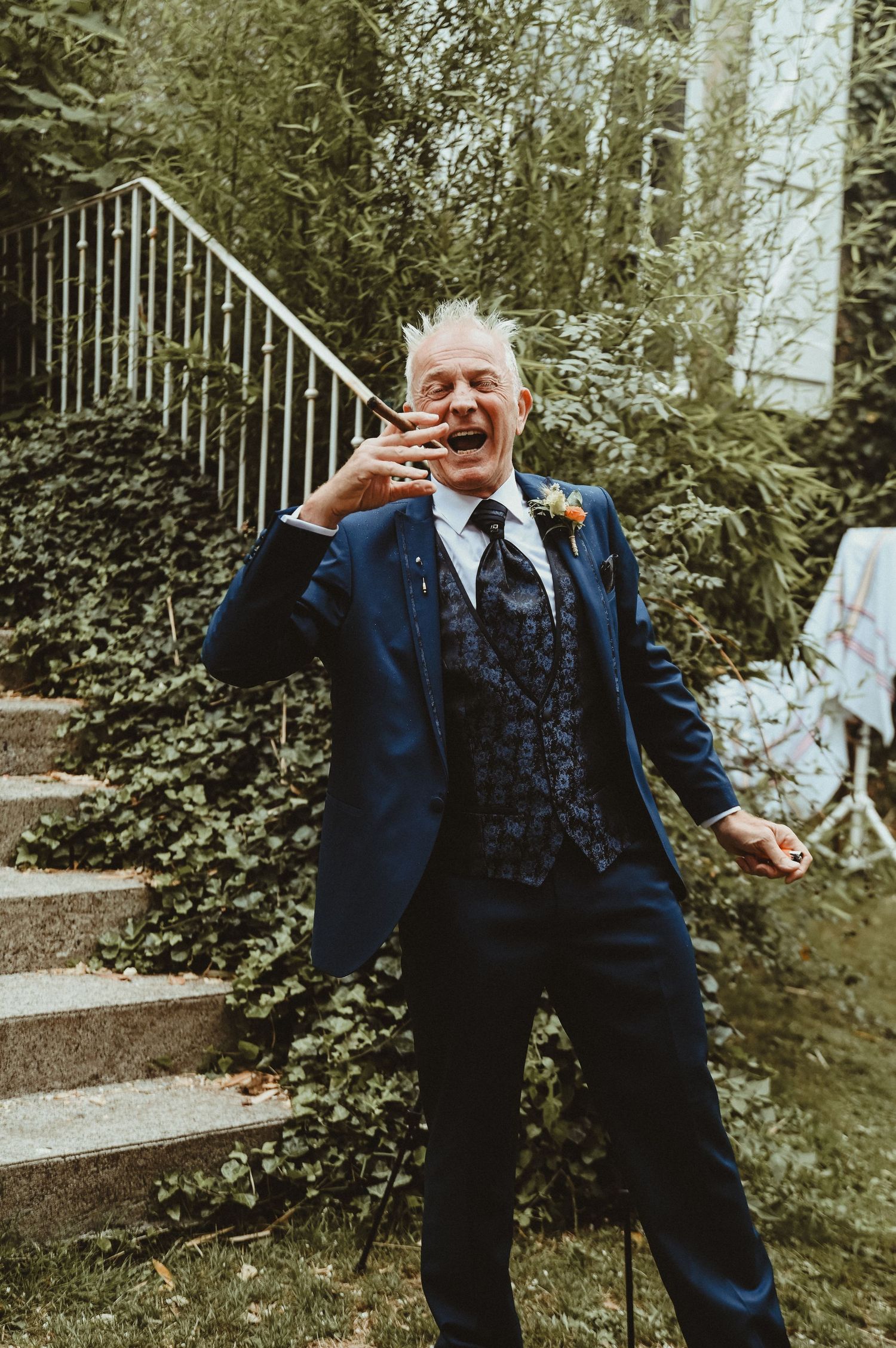 Wedding guest in a navy suit laughs expressively on outdoor stone steps.