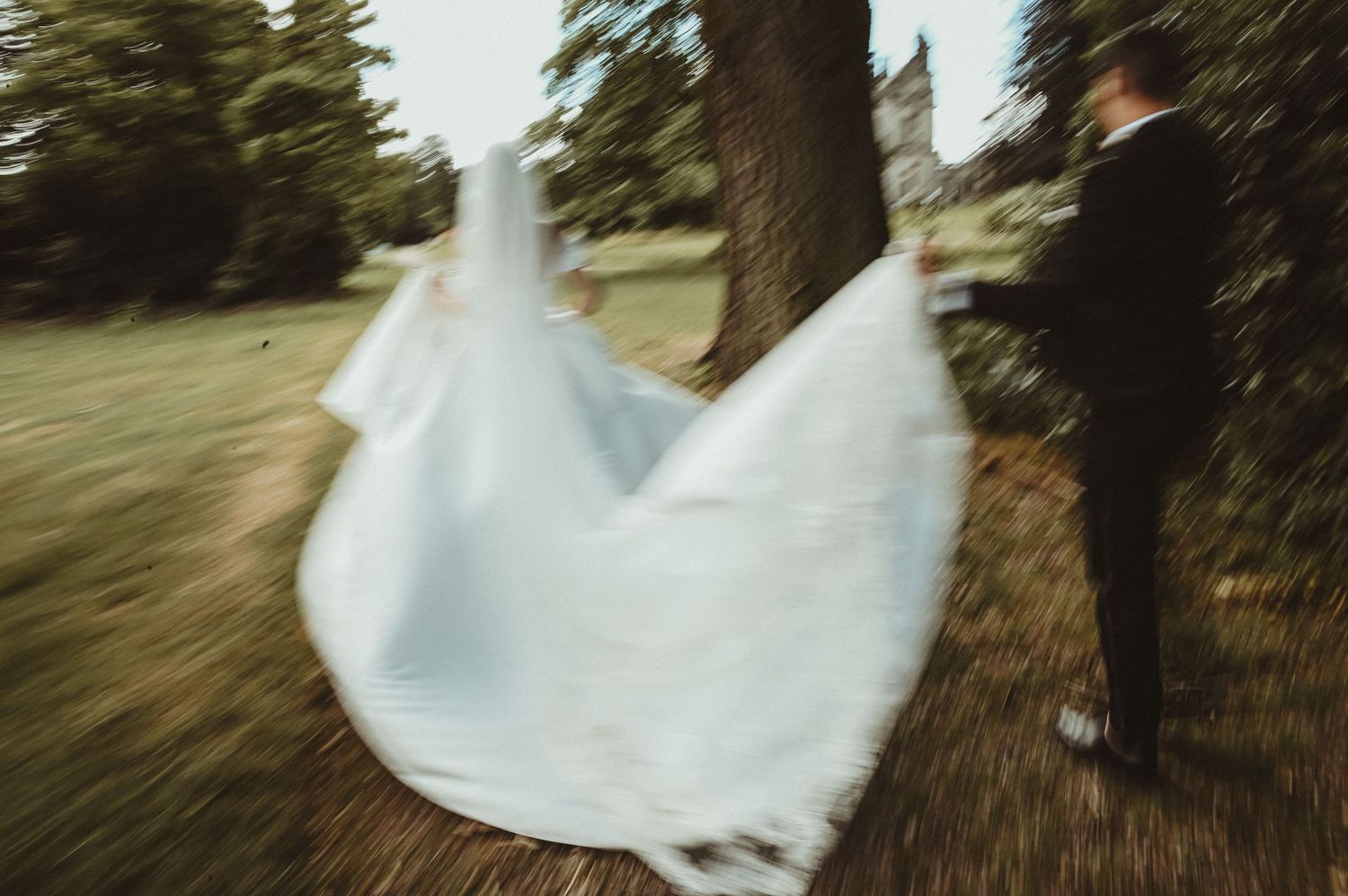A motion-blurred wedding dress flows dramatically against a natural outdoor setting with trees.