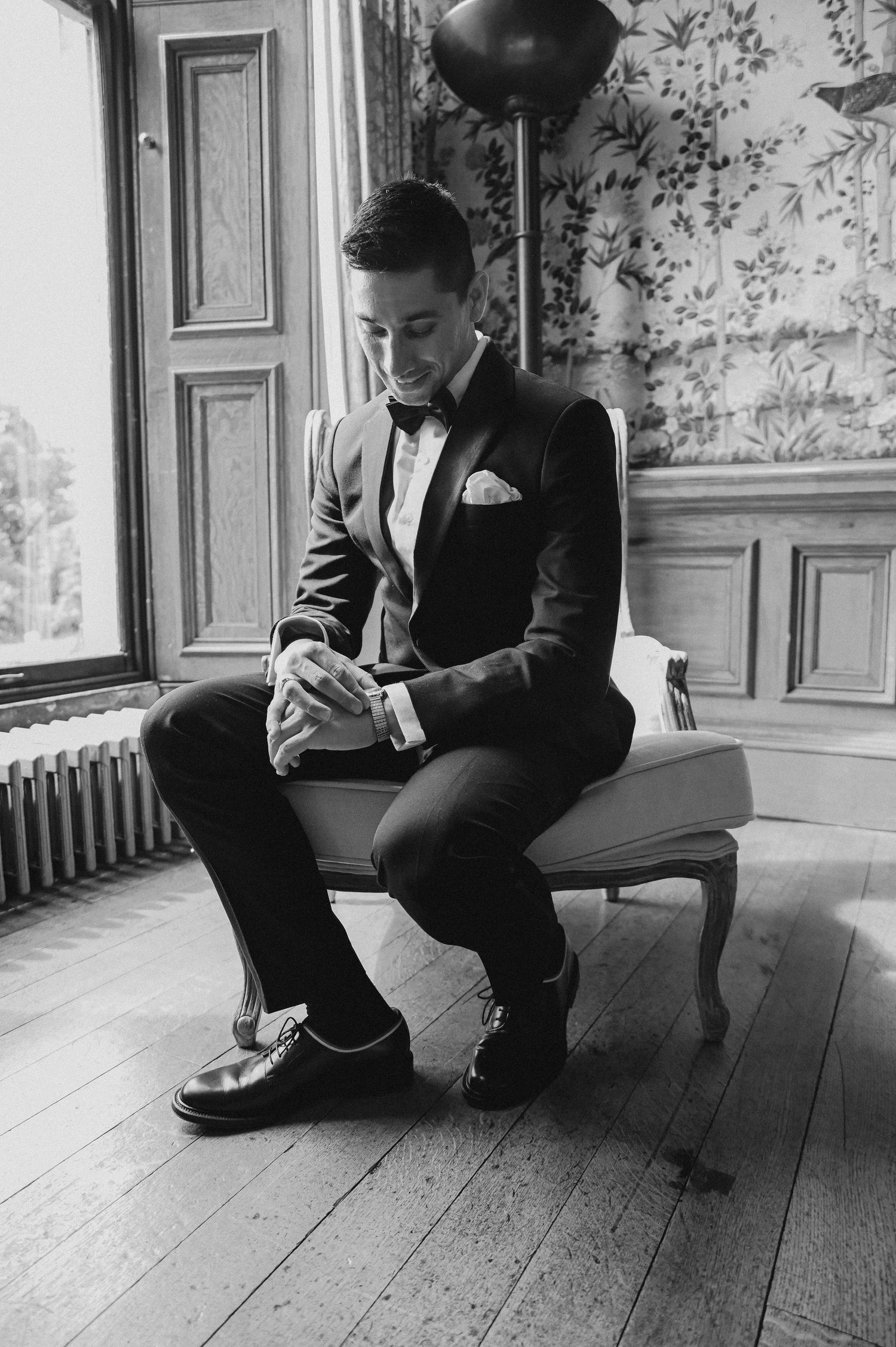 A suited figure seated on a chair in a room with floral wallpaper and hardwood floors.