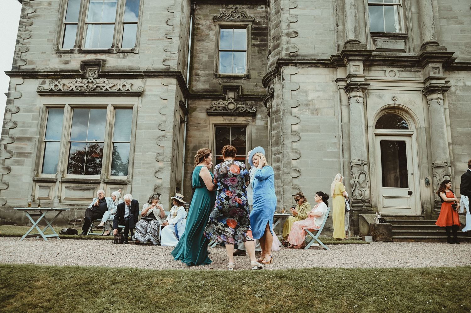 Guests in formal attire gather outside a grand stone mansion during an elegant outdoor event.