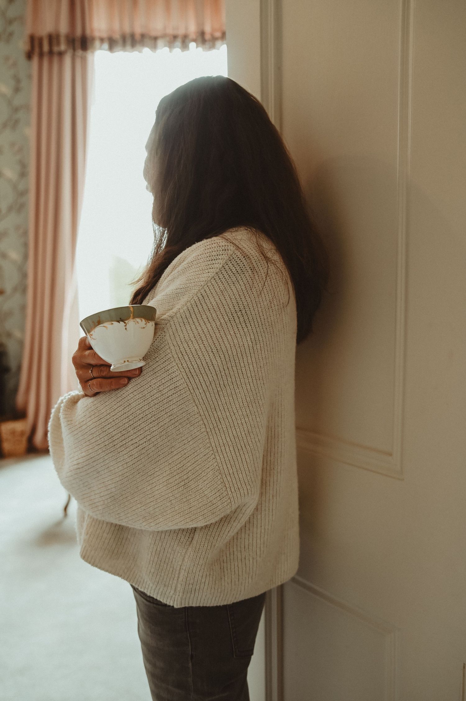 Silhouette of person in cozy sweater holding coffee cup by window with soft curtains.