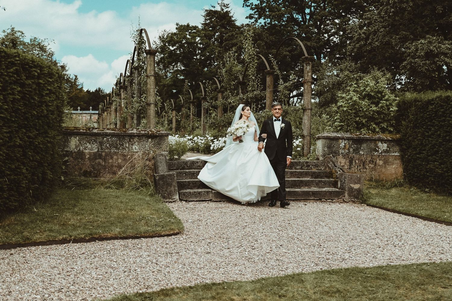 A couple in wedding attire poses on stone steps in a lush garden setting with classic architecture.