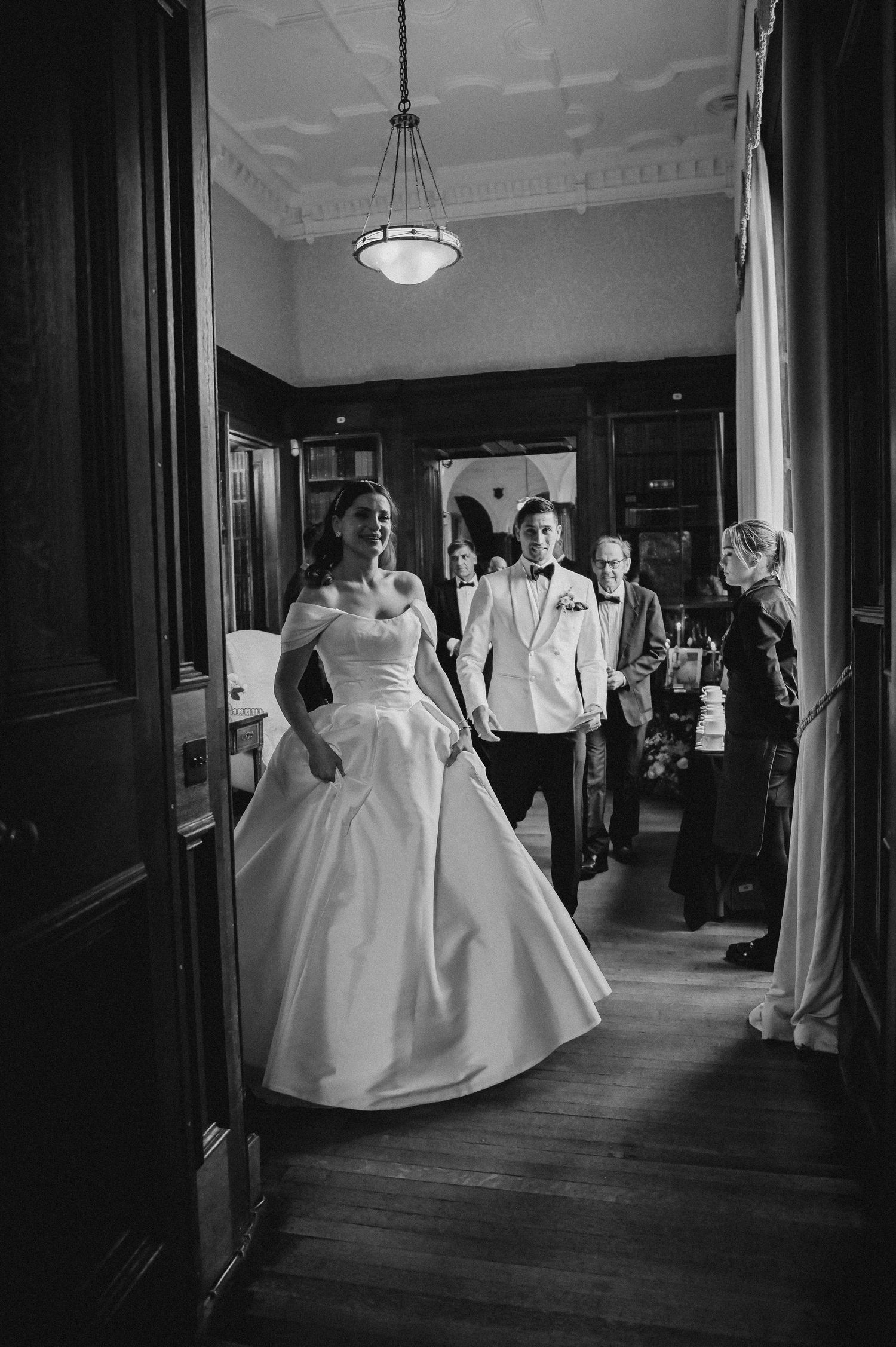 Black and white photo of elegant bridal party walking through a grand hallway with wooden details.
