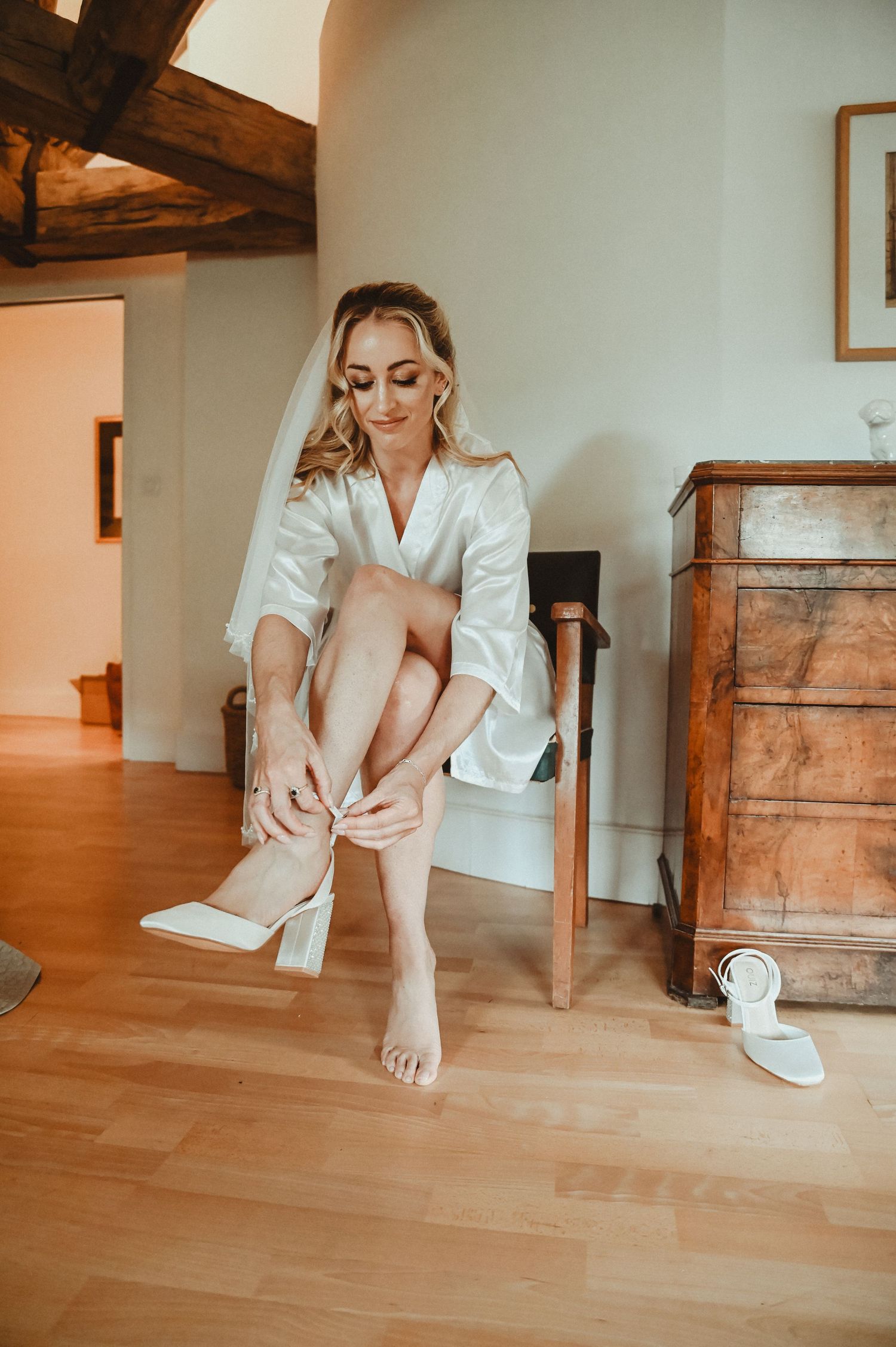 Person in a white silk robe putting on white heels while getting ready on a wooden floor.