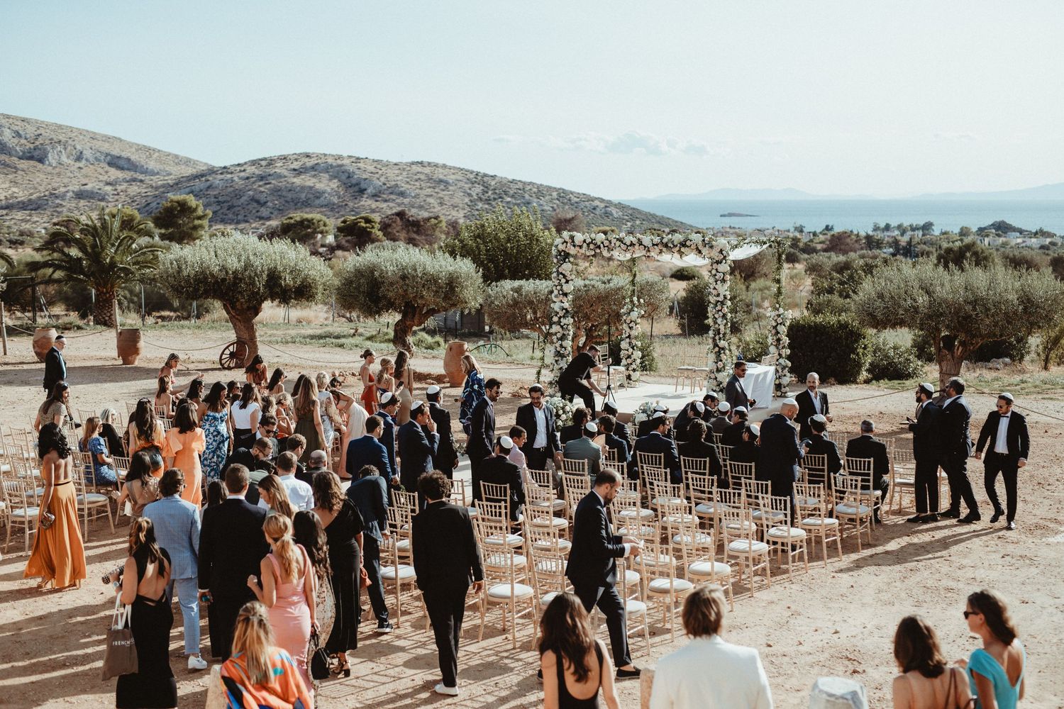 Wedding guests gather around white chairs set up for an outdoor ceremony with mountain views.