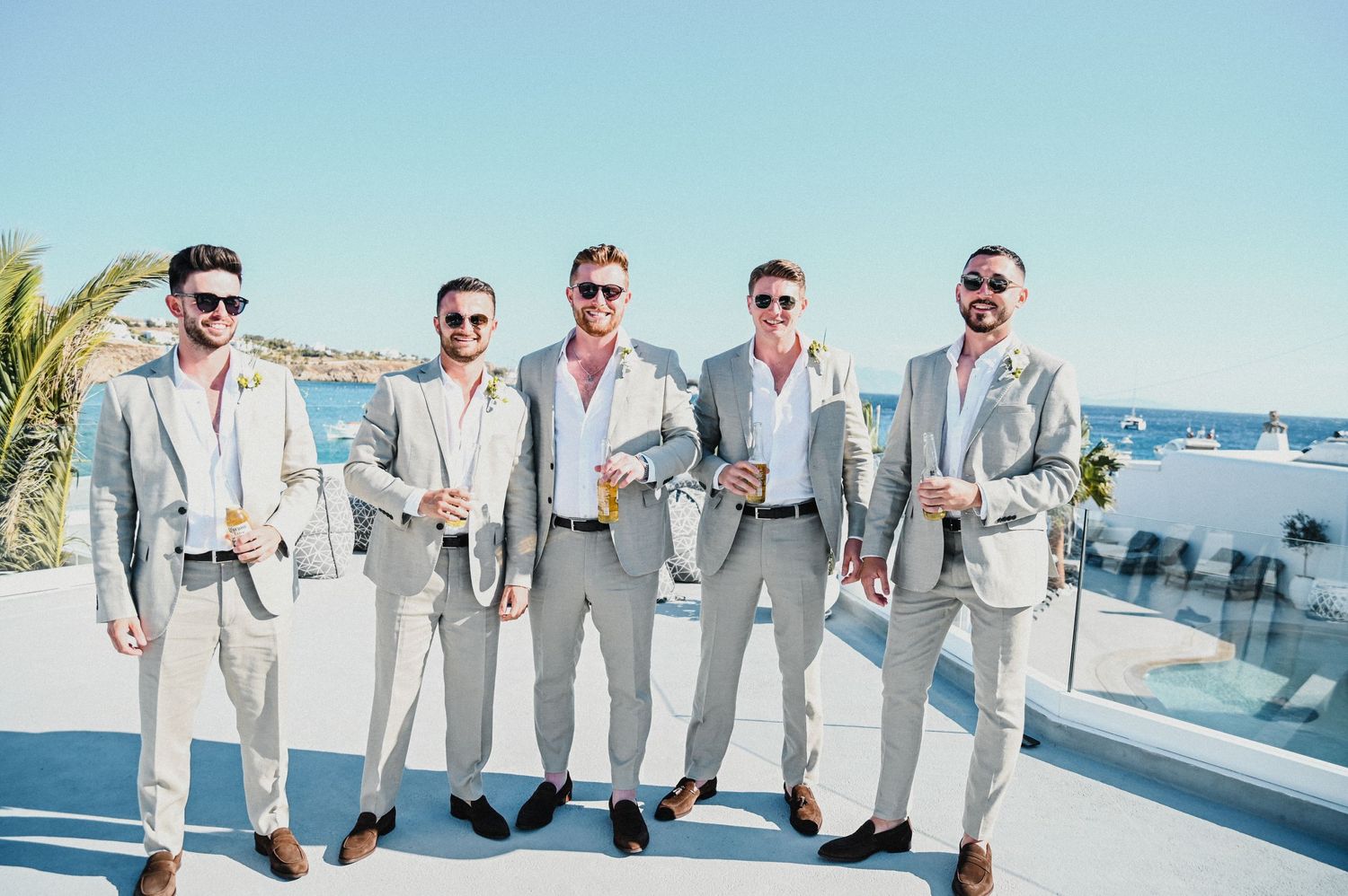 Five groomsmen in coordinated gray suits pose with drinks on a sunny terrace.