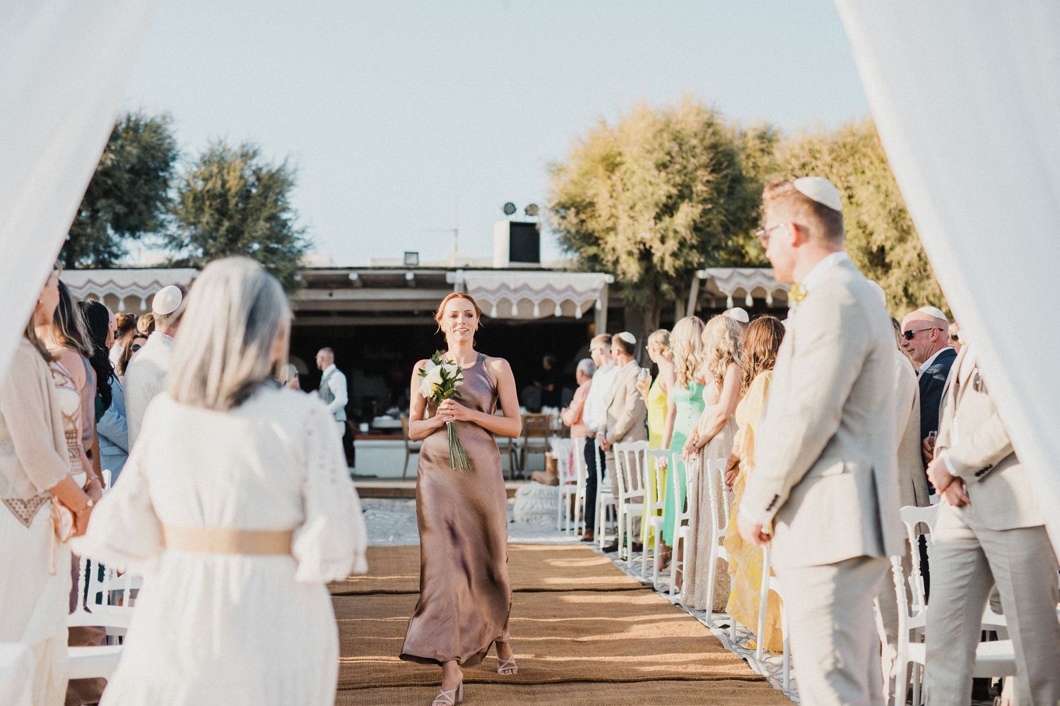 Wedding ceremony taking place on a wooden deck with guests seated in white chairs in natural sunlight.