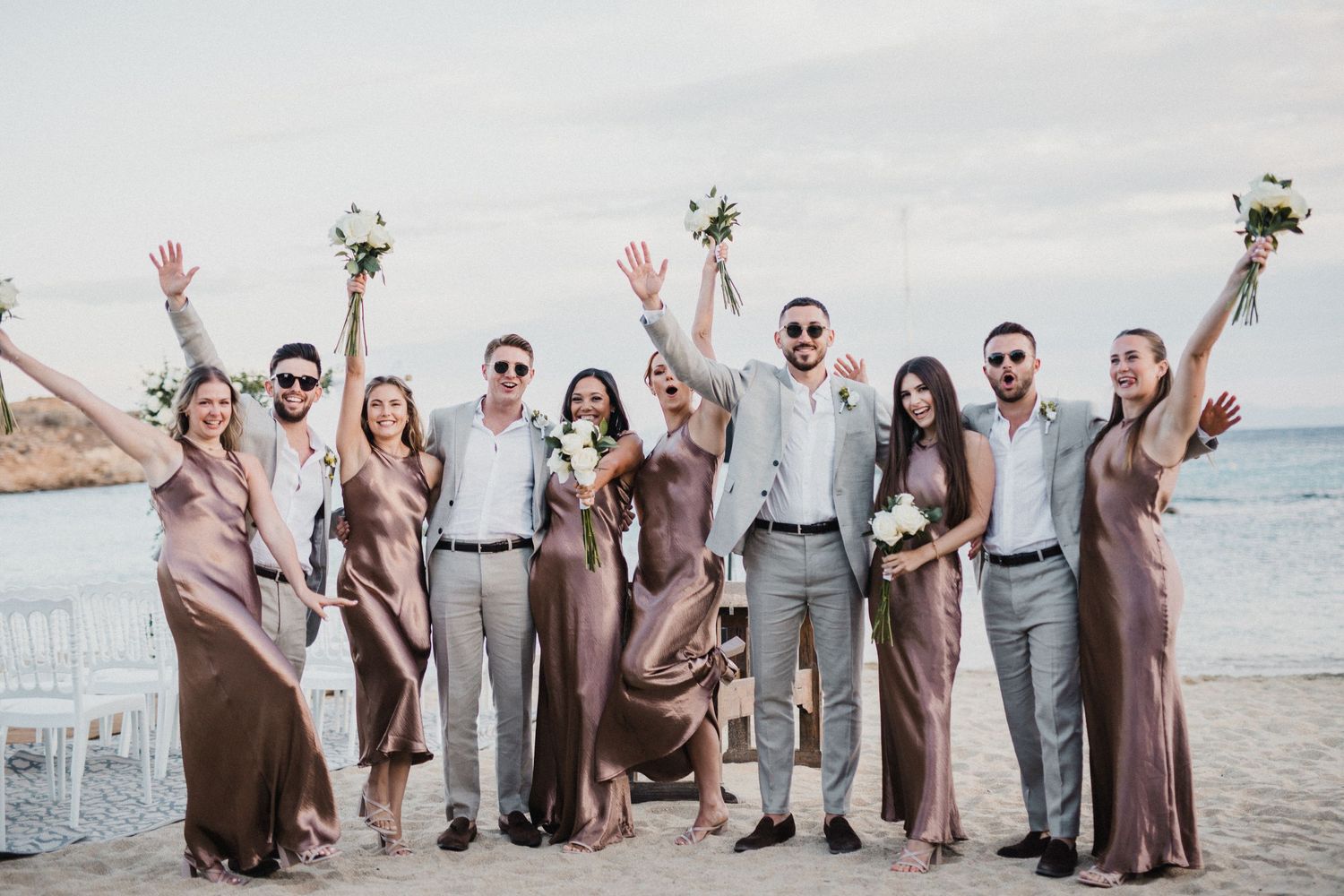 Wedding party celebrates on beach with raised bouquets in neutral-toned attire.
