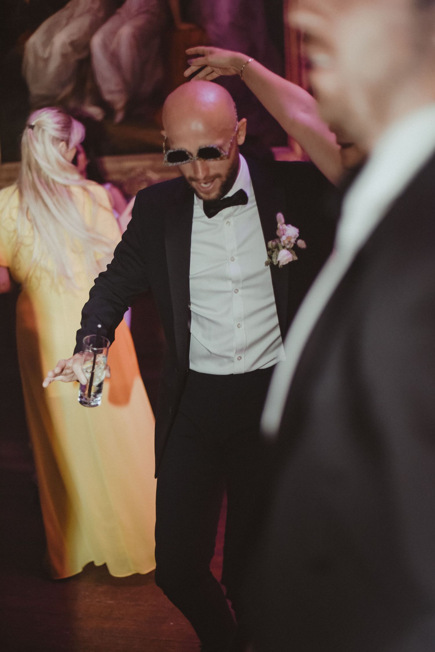 A guest dances enthusiastically at a wedding reception wearing a tuxedo and boutonniere.