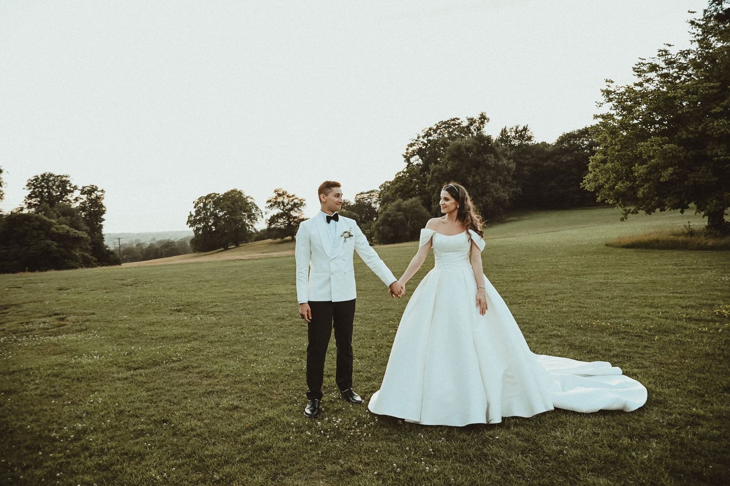 A couple poses together on a grassy hillside in formal wedding attire.