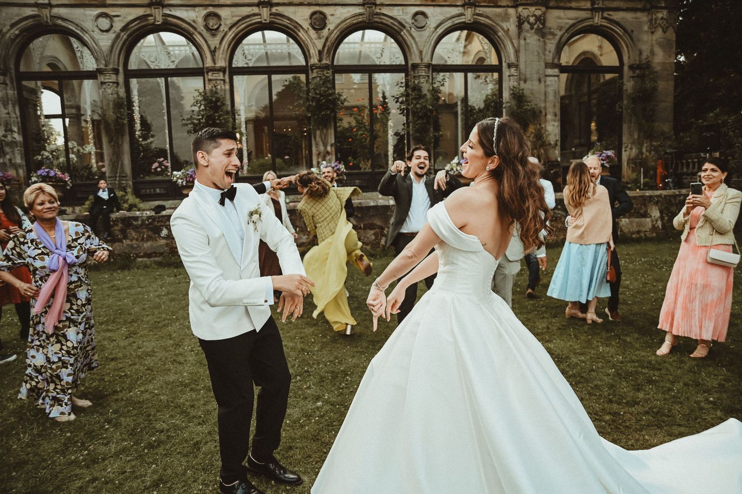 Newlyweds dance enthusiastically on the lawn of a historic venue with arched windows.
