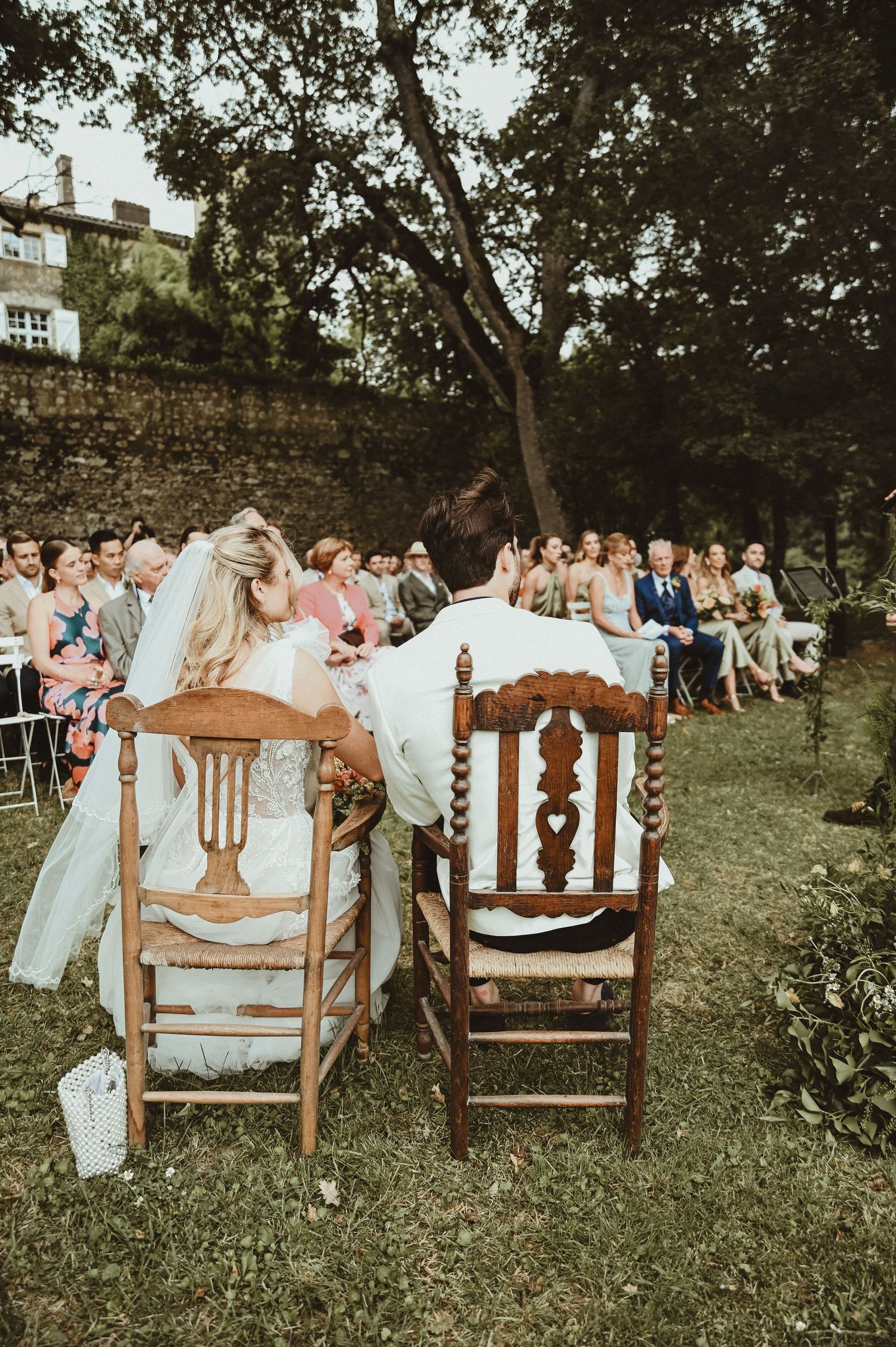 A couple sits in wooden chairs during an outdoor garden wedding ceremony with guests seated behind them.