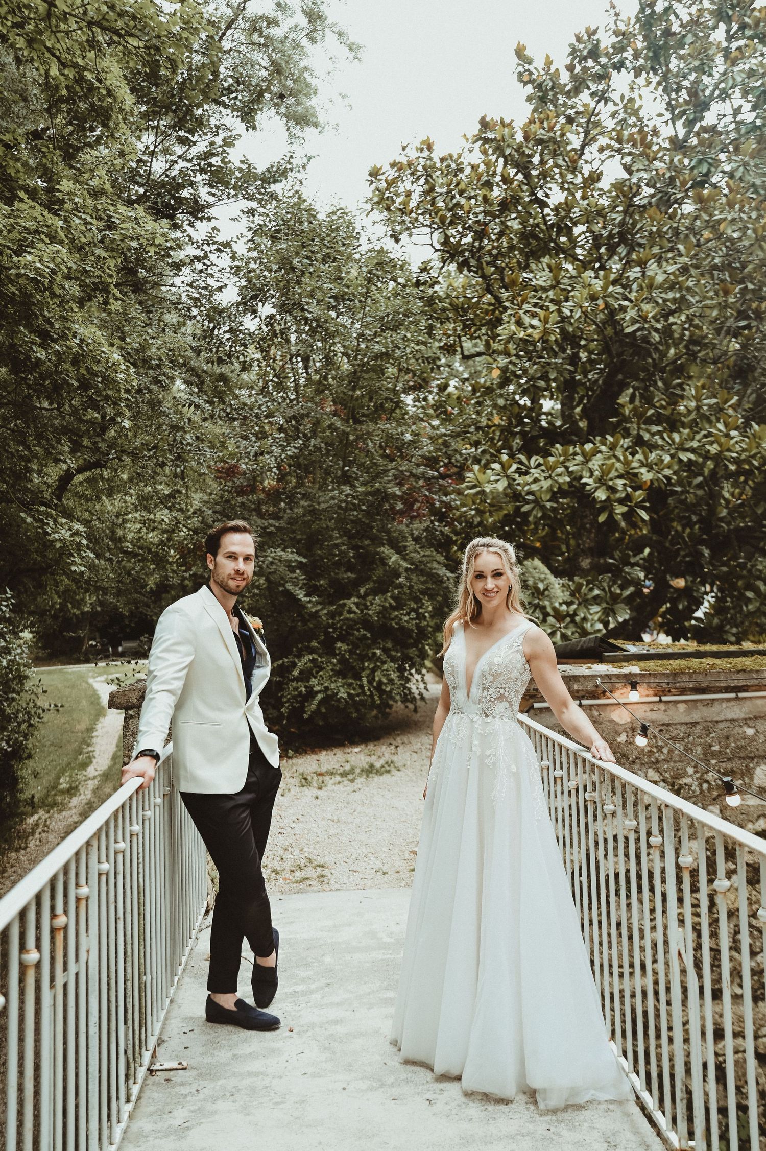 A bride and groom pose on opposite sides of a white metal bridge in a garden setting.