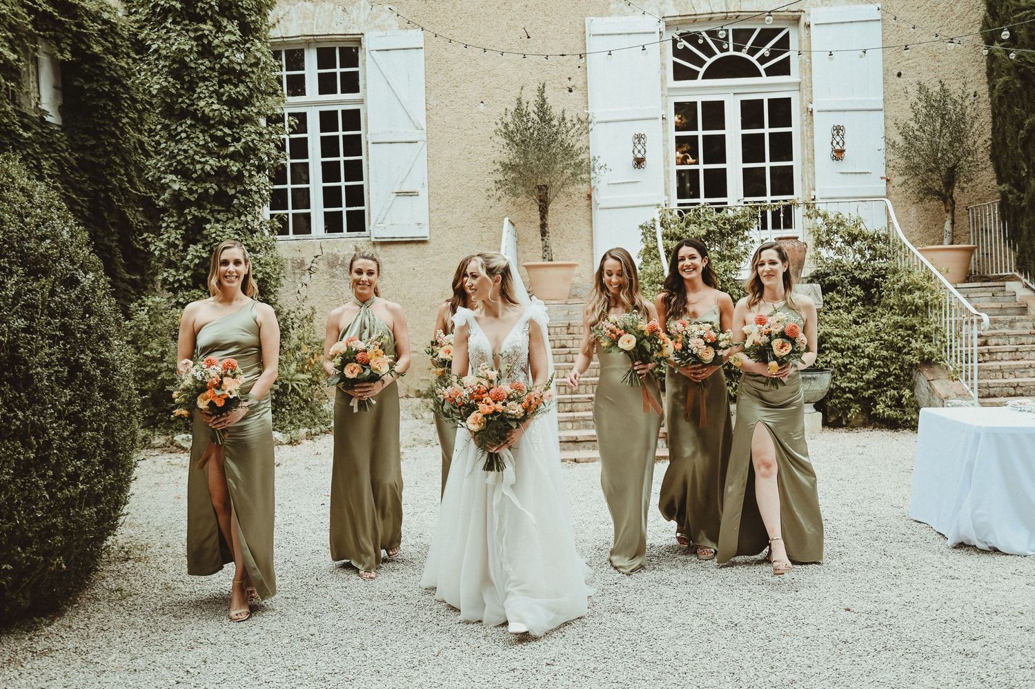 Bridal party poses outside a French chateau with peach bouquets and sage colored dresses.