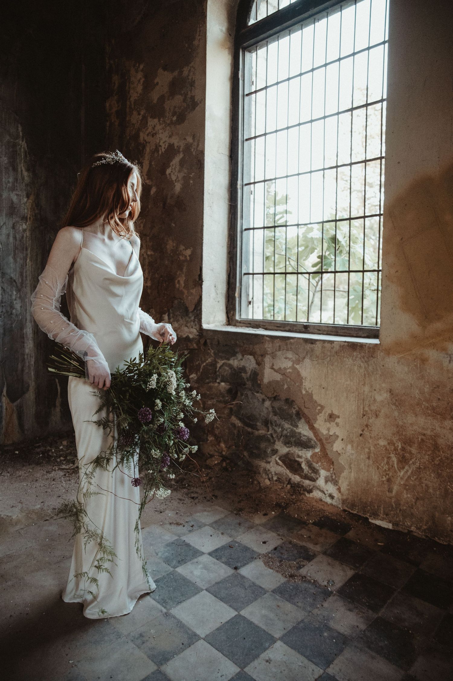 Elegant bride in flowing dress holding bouquet by vintage window with checkered floor.