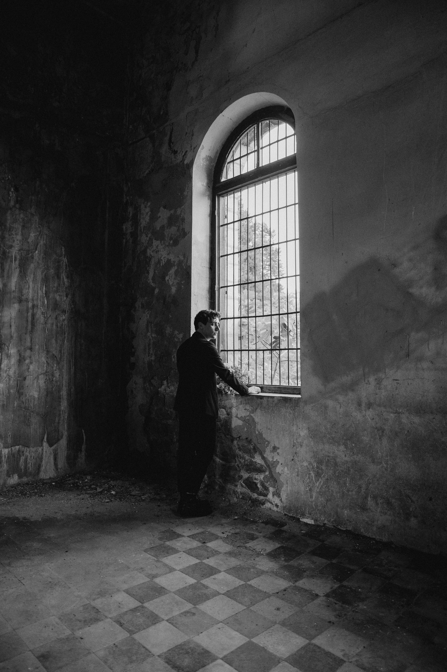 A silhouetted figure stands at a large arched window in a dimly lit room with checkered floor tiles.