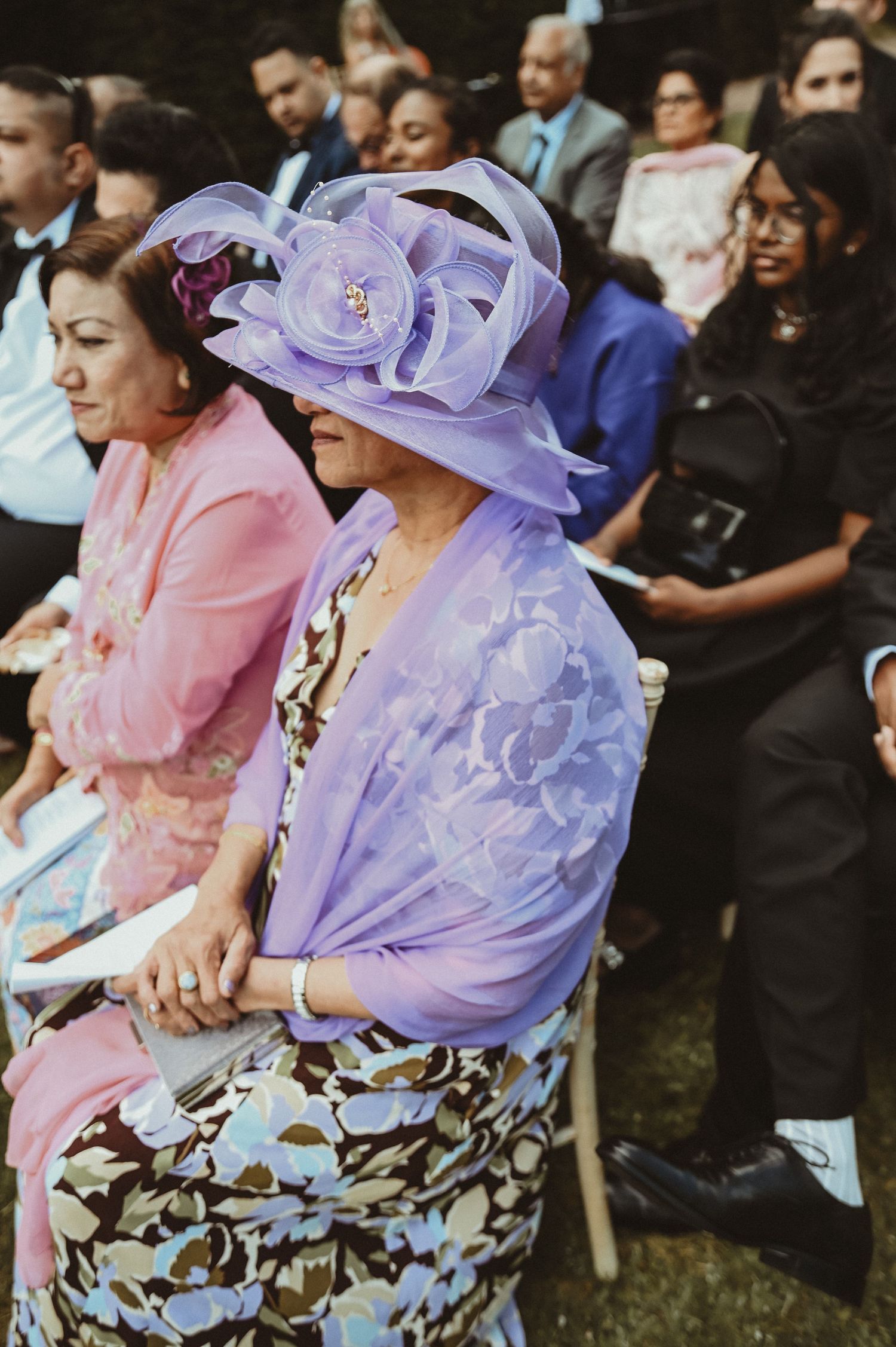 Guests at a wedding wearing elegant pastel outfits including a striking purple hat with floral detail.