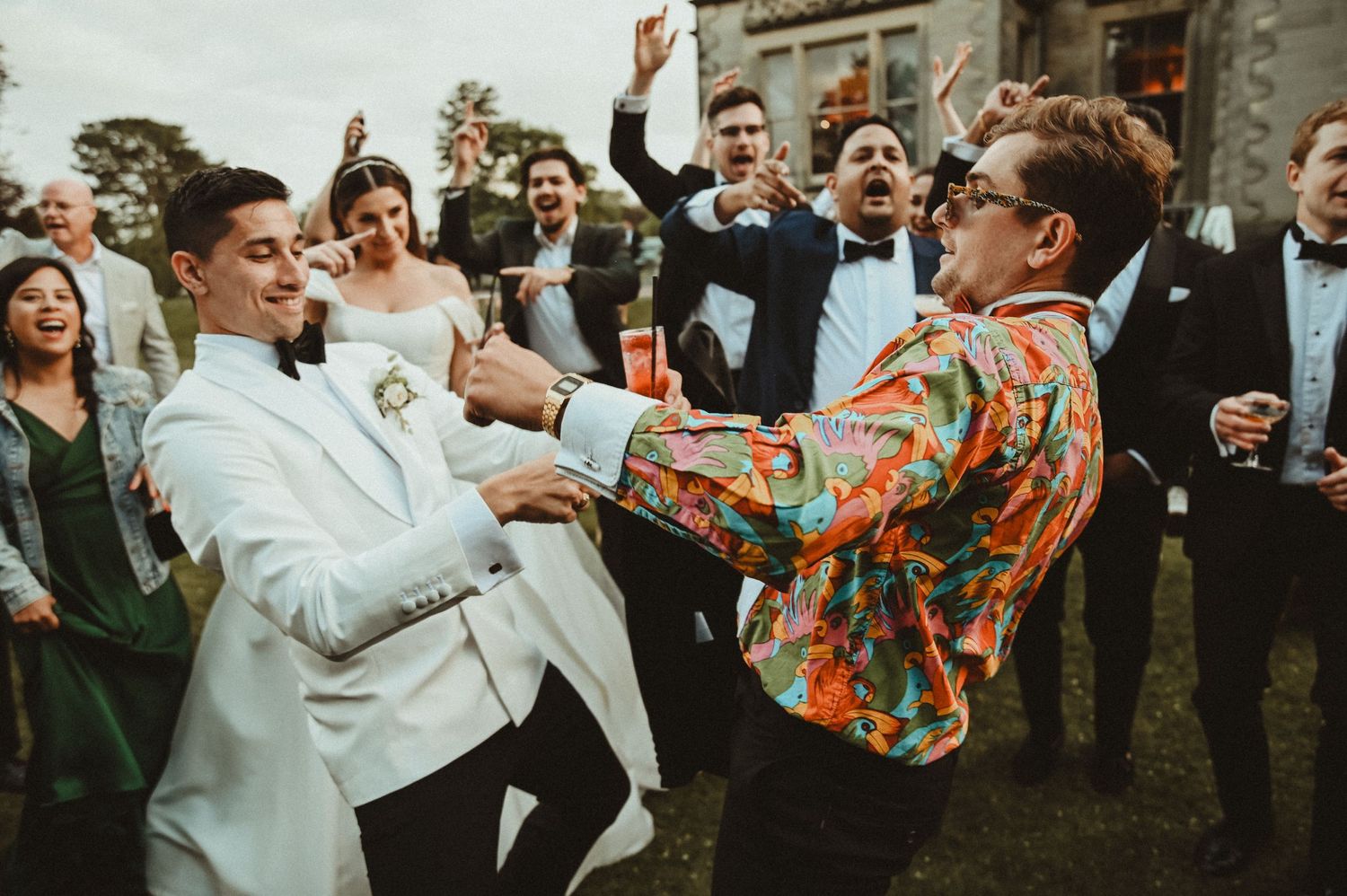 Wedding guests in formal attire dance energetically at an outdoor reception.