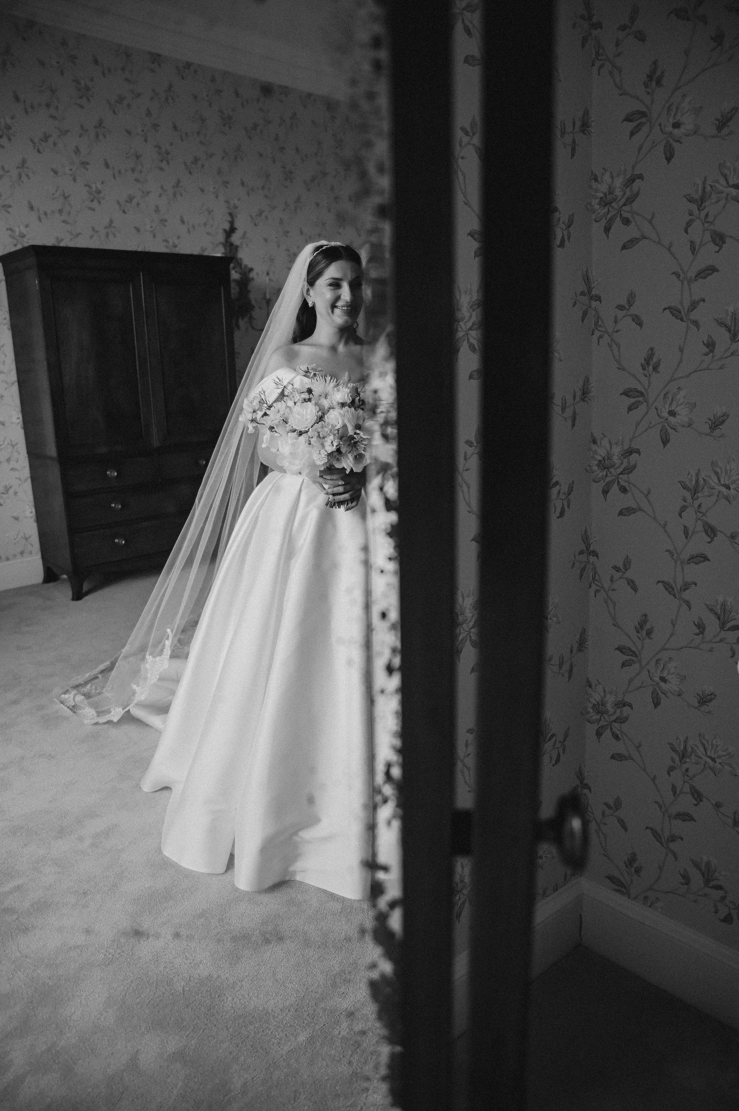 Black and white photograph of a wedding dress and bouquet glimpsed through a doorway with floral wallpaper.