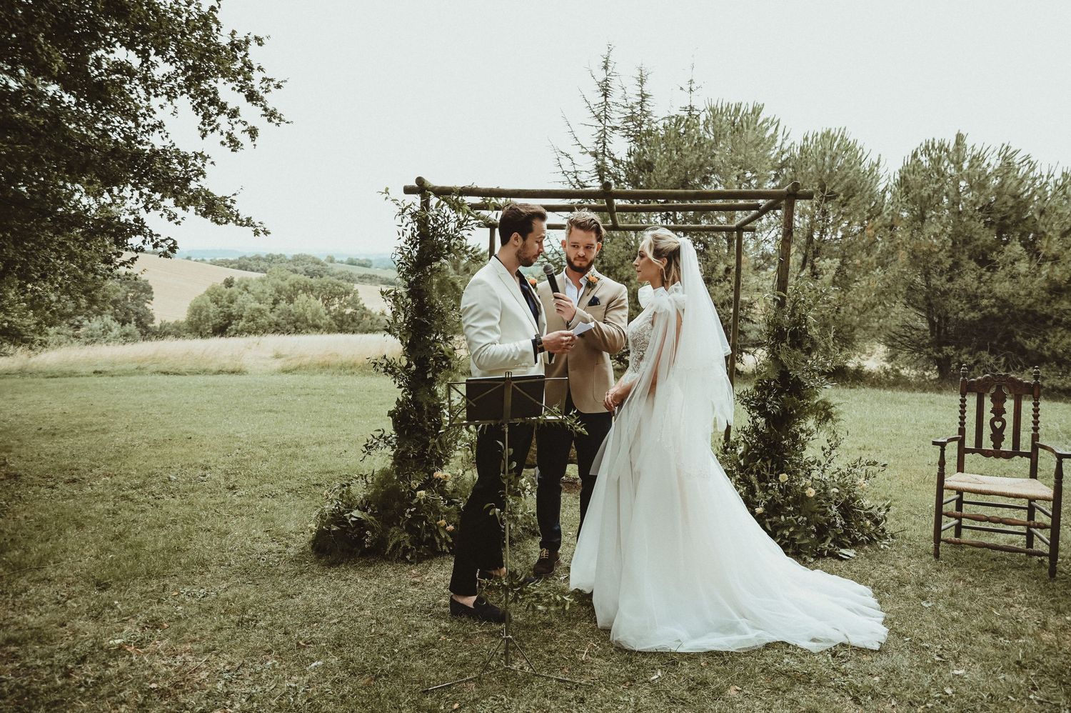 A wedding ceremony takes place under a rustic wooden pergola decorated with greenery in an outdoor garden setting.