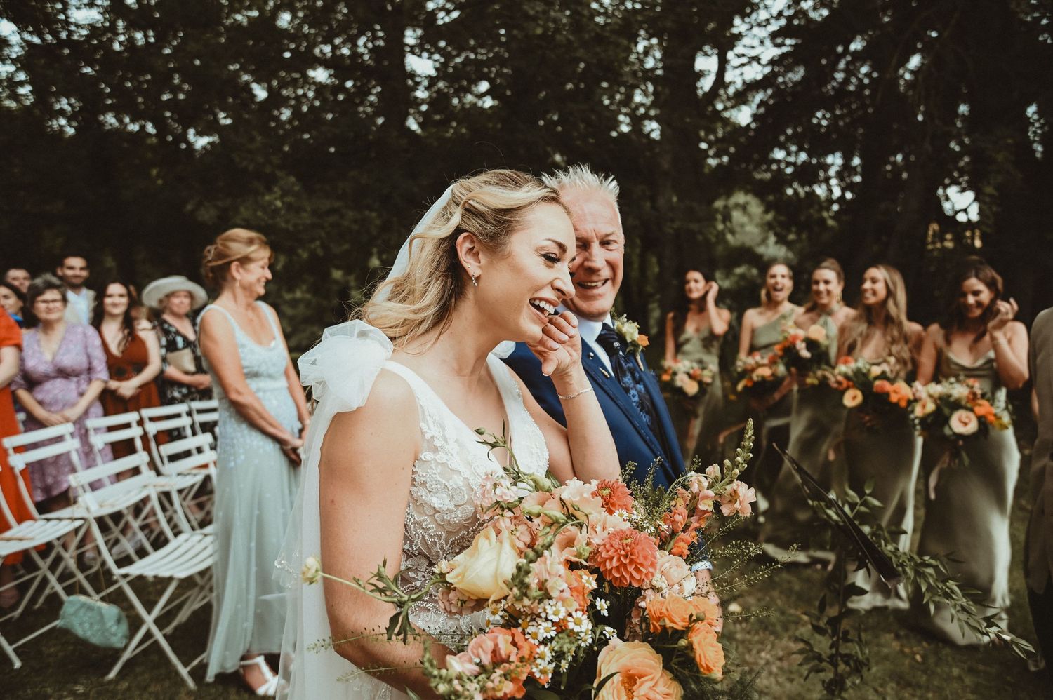 Wedding party celebrates during outdoor ceremony with white chairs and floral decorations.