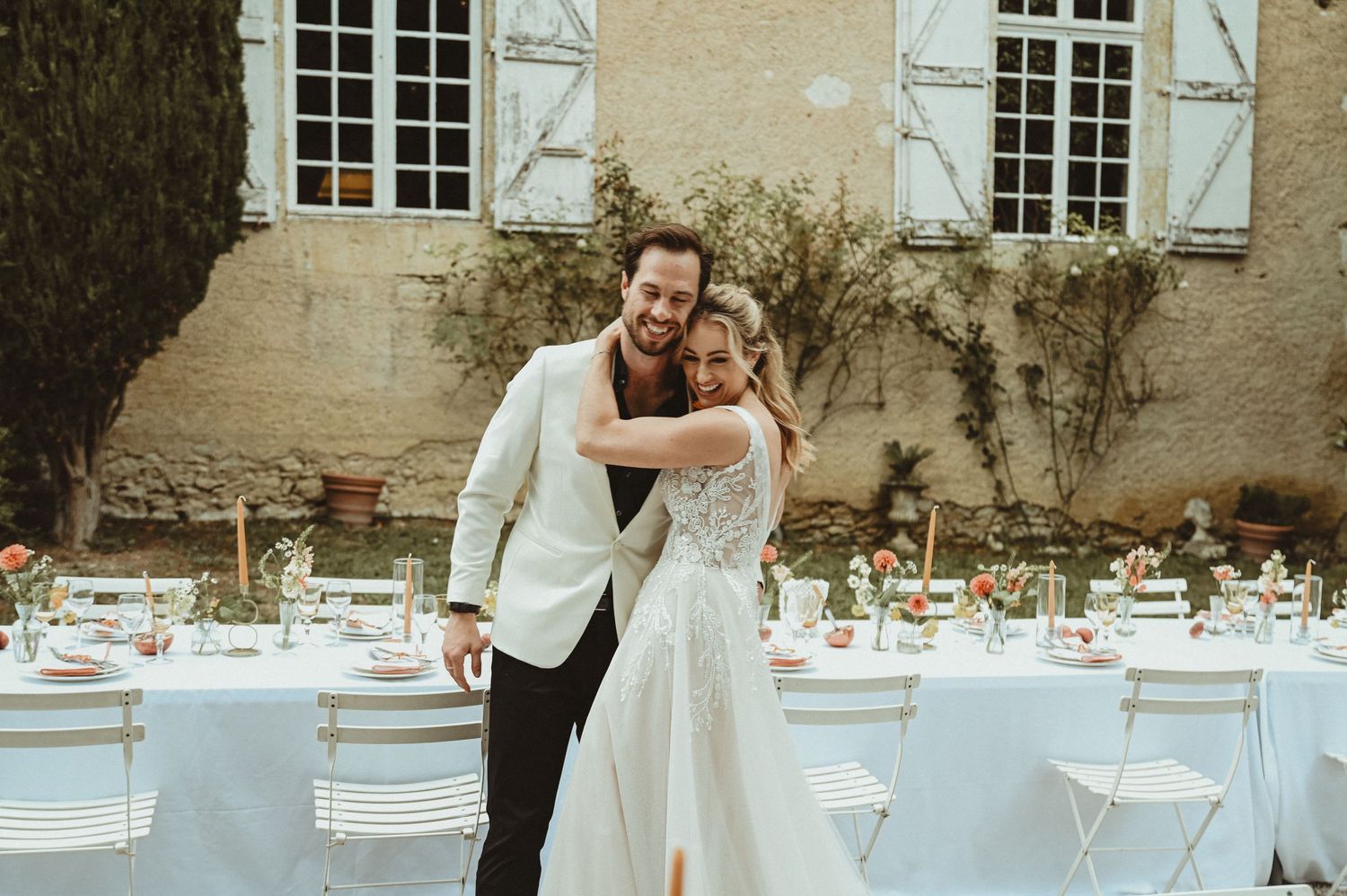 Wedding celebration setup with elegant table settings and white chairs against a rustic stone building.