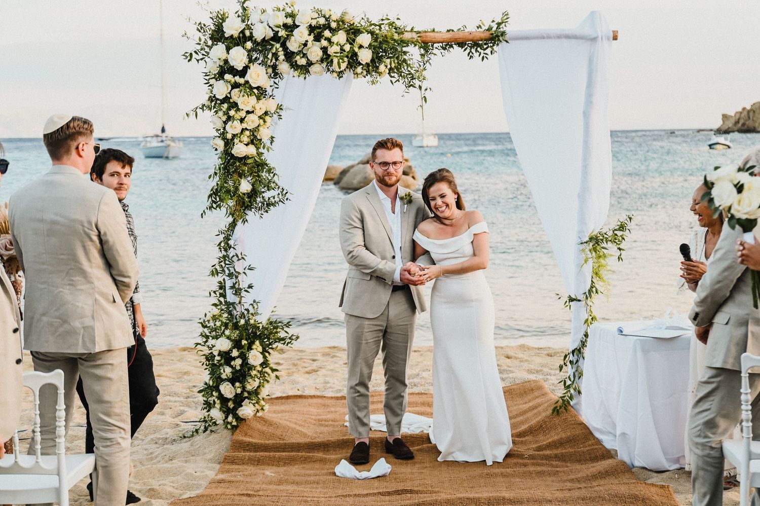 Wedding ceremony on beach with decorated arch and guests watching couple.