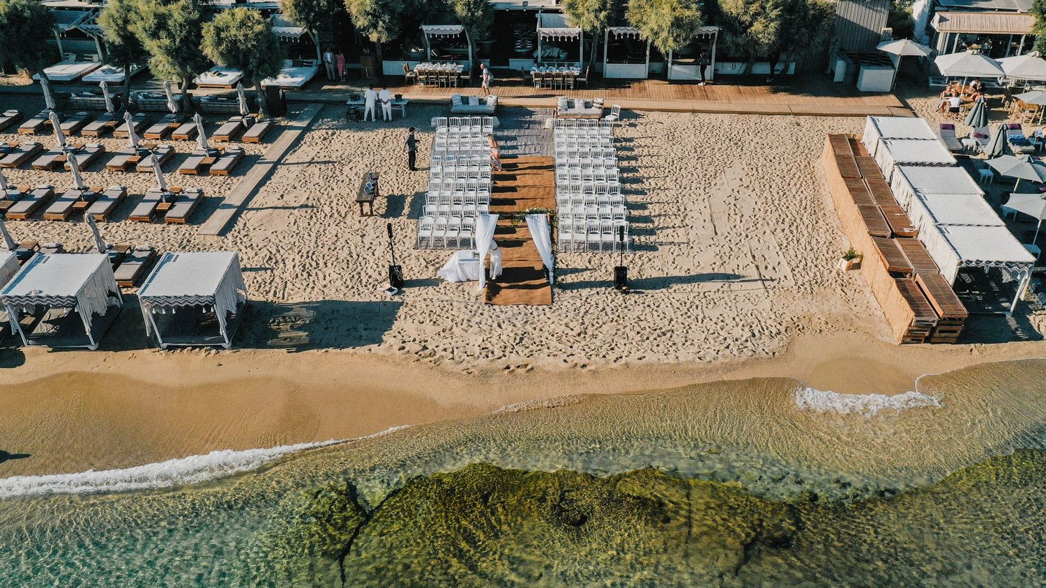 Birds-eye view of a coastal beach club with neat rows of sun loungers and umbrellas on sandy shore.