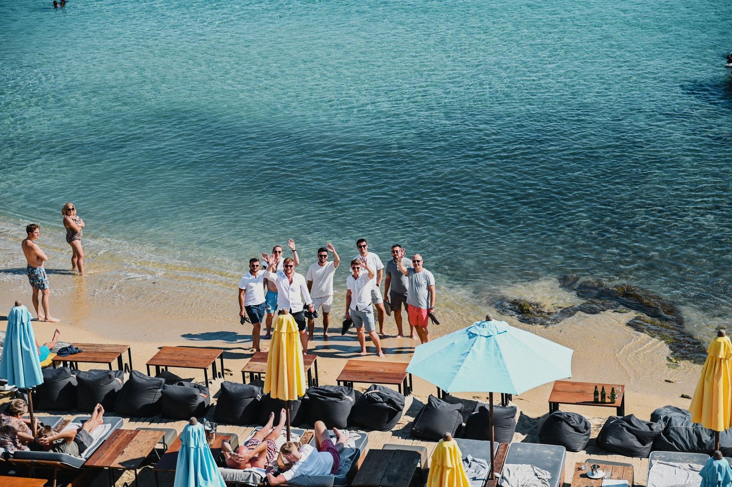 Wedding guests gather at a beachside venue with turquoise water and blue umbrellas on a sunny day.