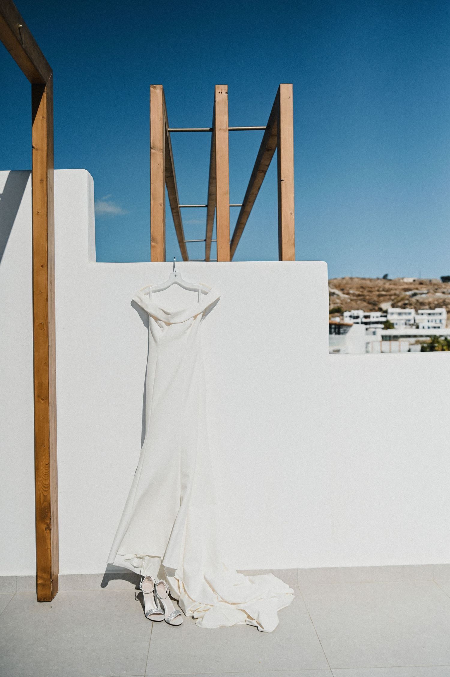 White wedding dress hanging against a minimalist white wall with wooden beams under blue sky.