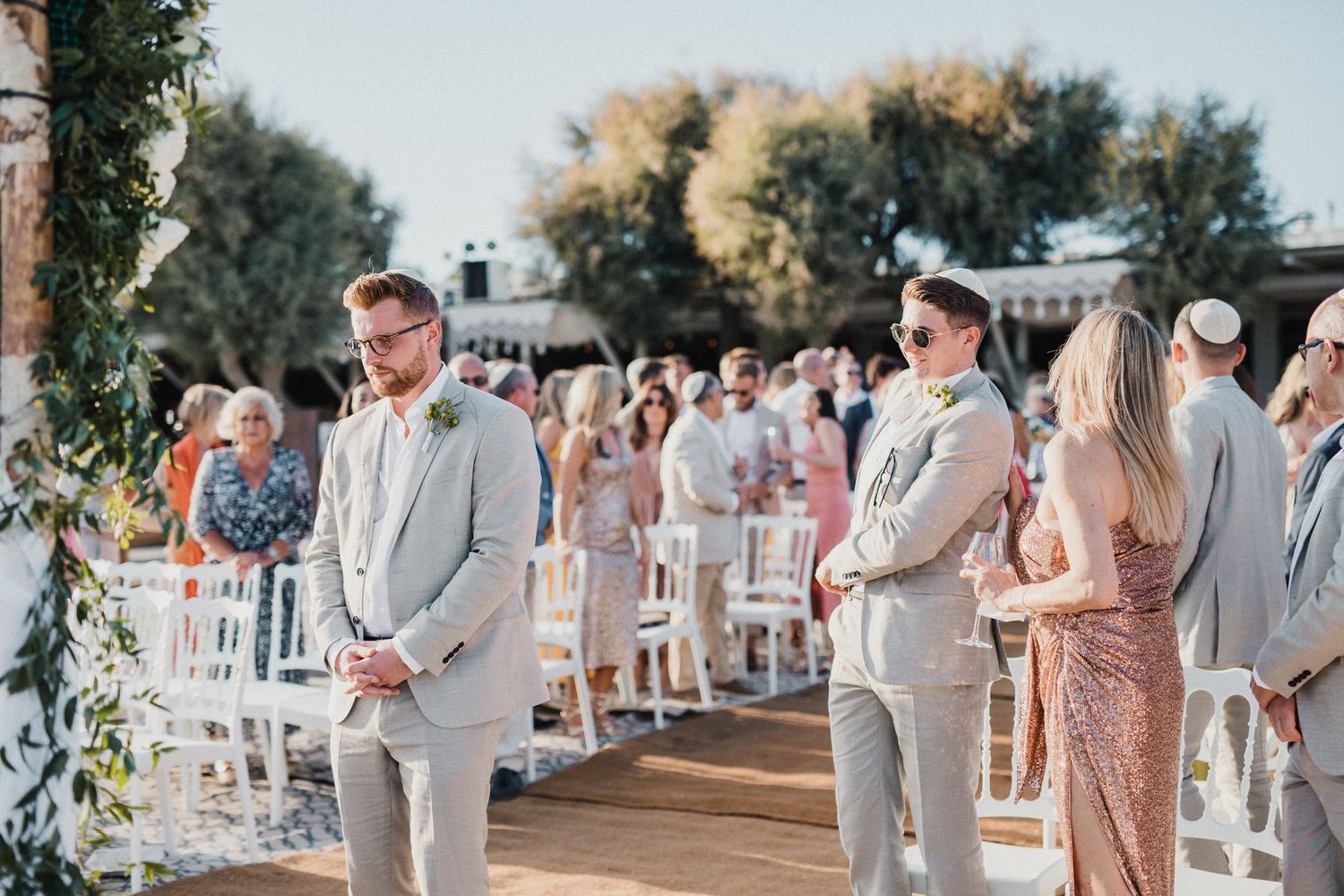 Outdoor wedding ceremony setup with guests in light colored attire standing on a wooden platform.