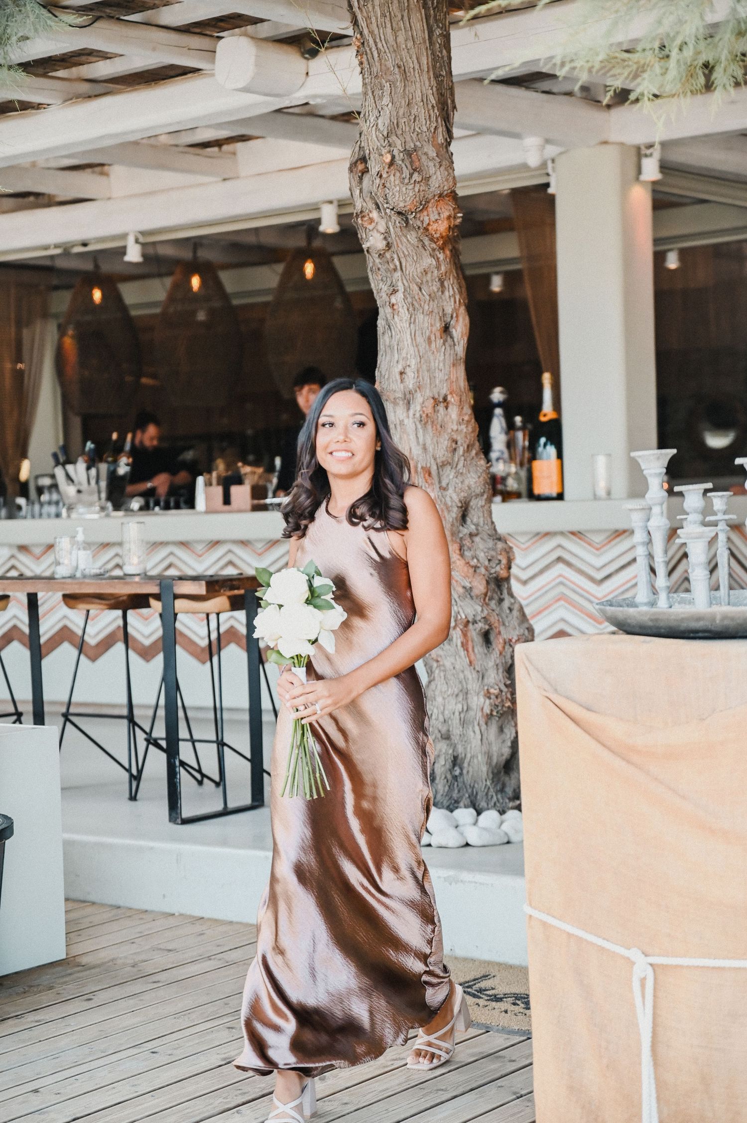 Wedding party member in silk champagne dress poses with bouquet in modern coastal venue.