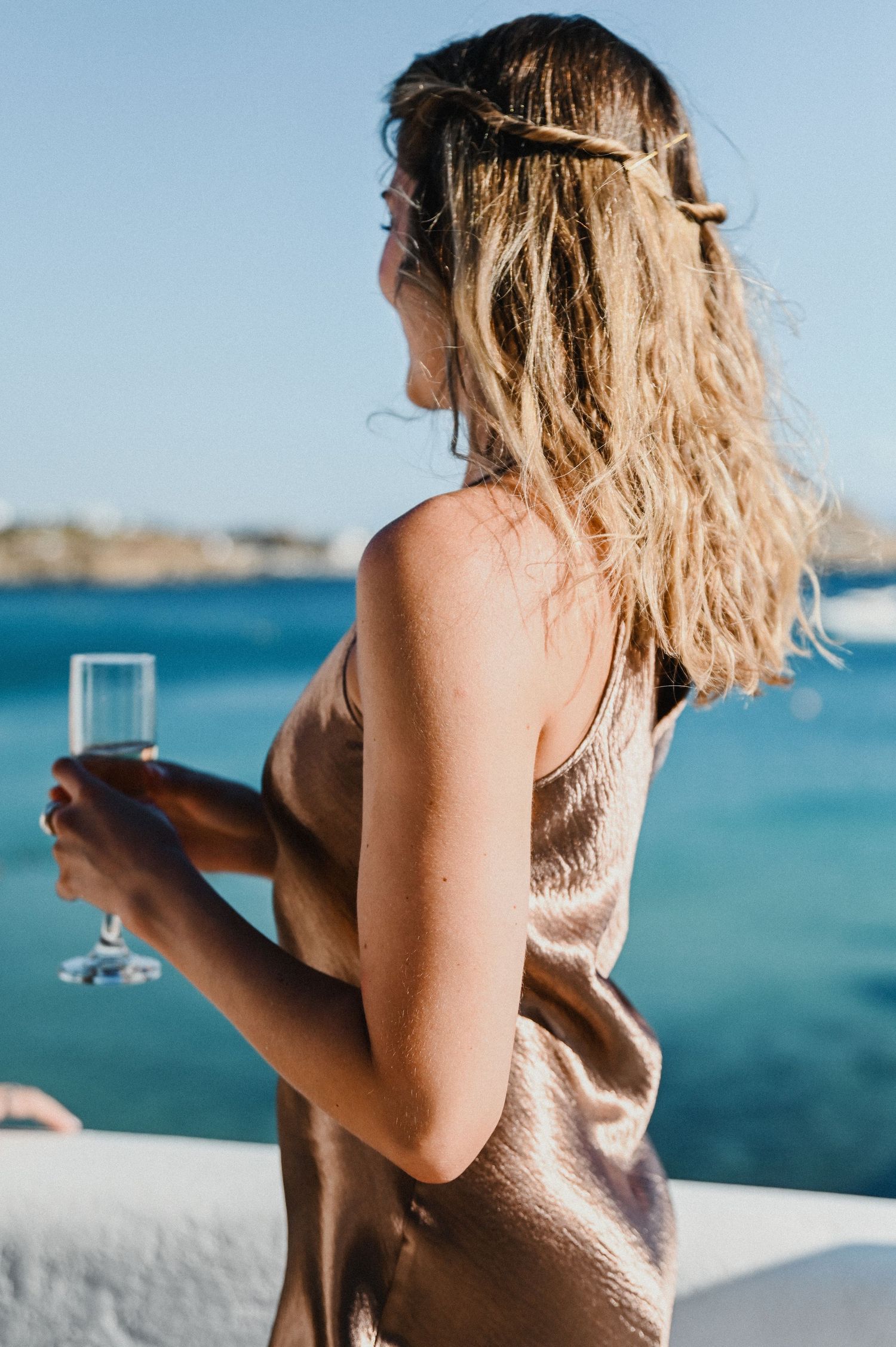 Woman in champagne dress holds champagne glass on coastal balcony with ocean view.