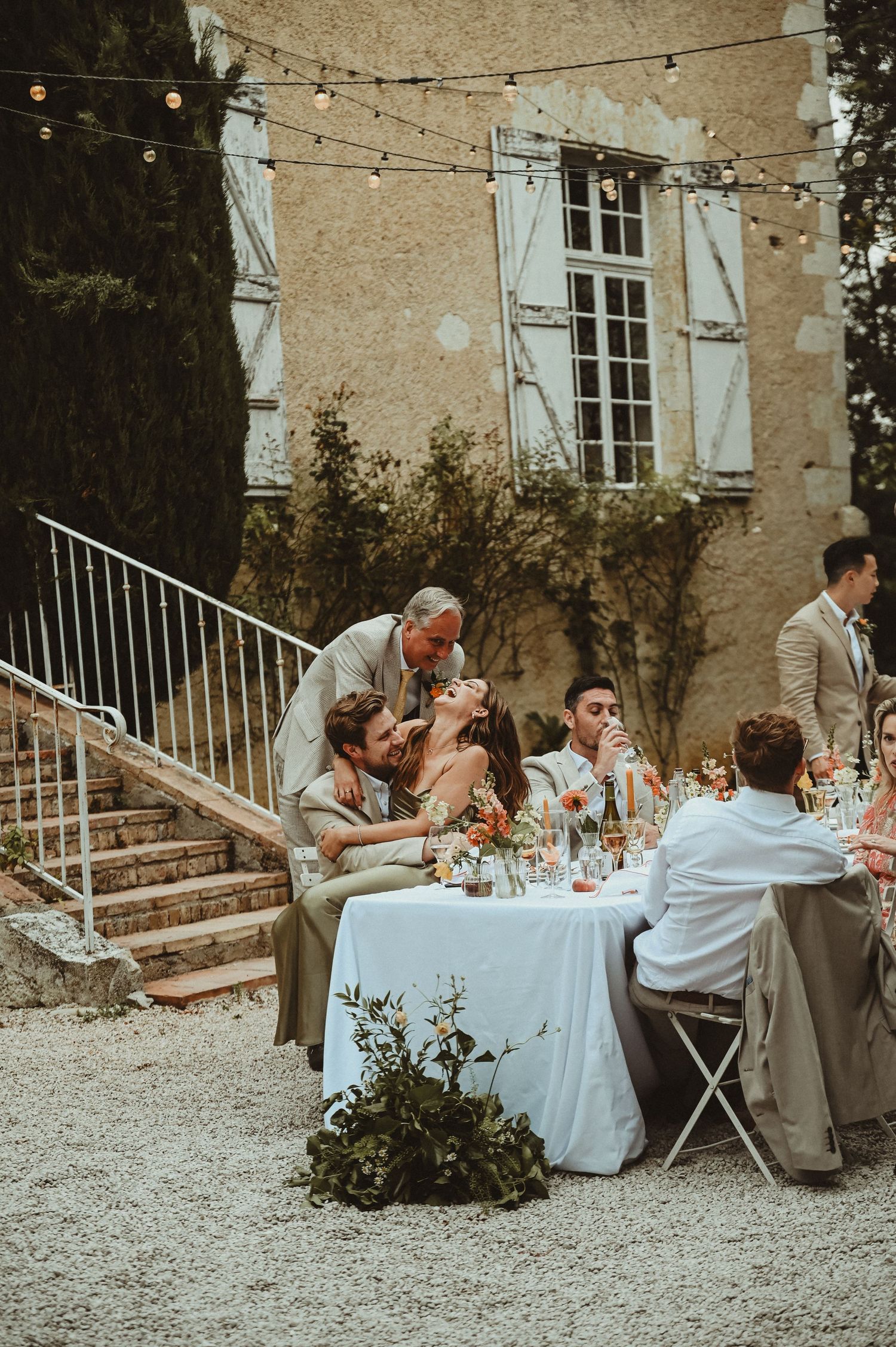 Guests dining at elegant outdoor wedding reception beside stone chateau with string lights.