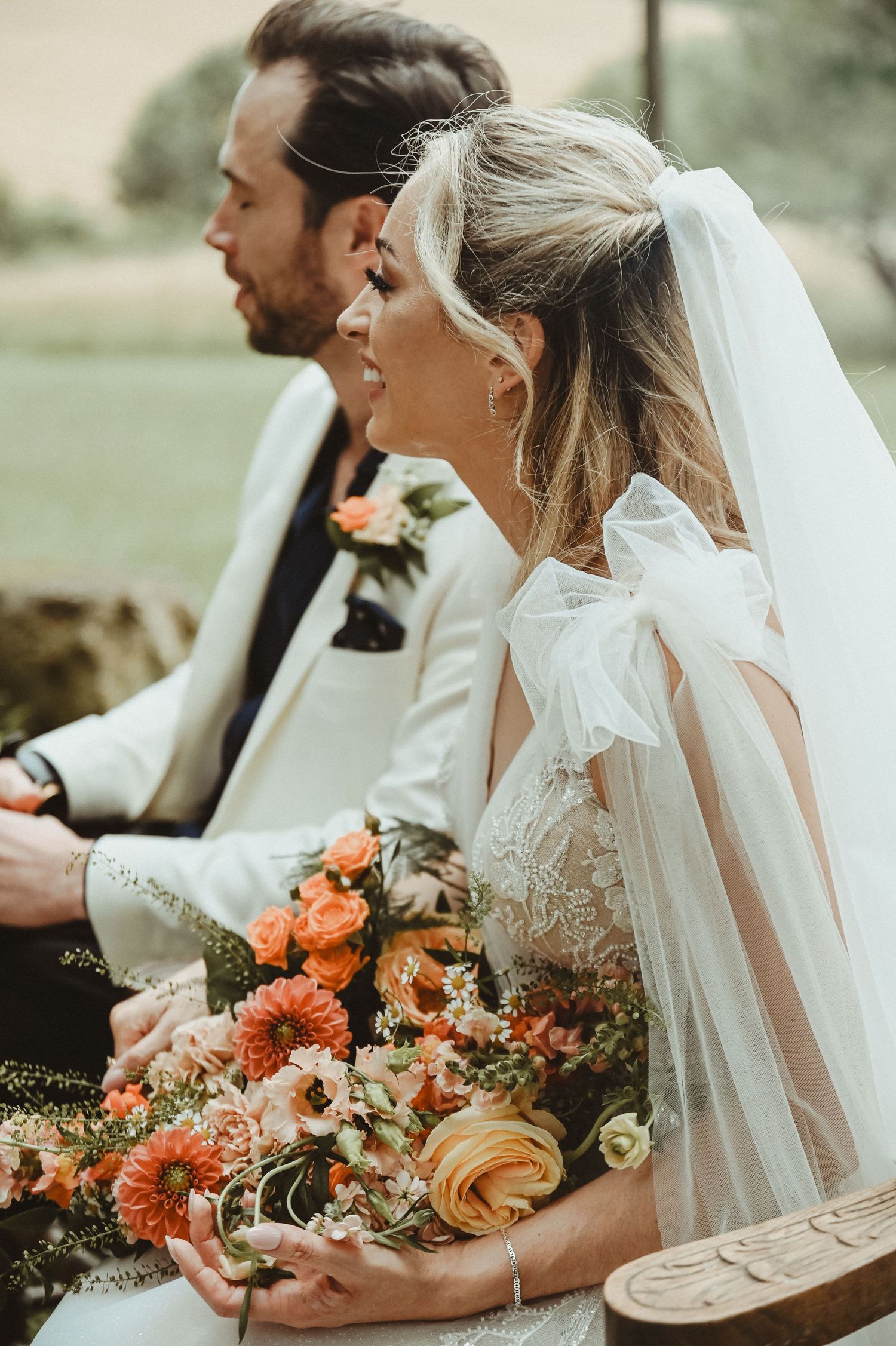 A bride in white holds a coral and peach bouquet during an outdoor wedding ceremony.