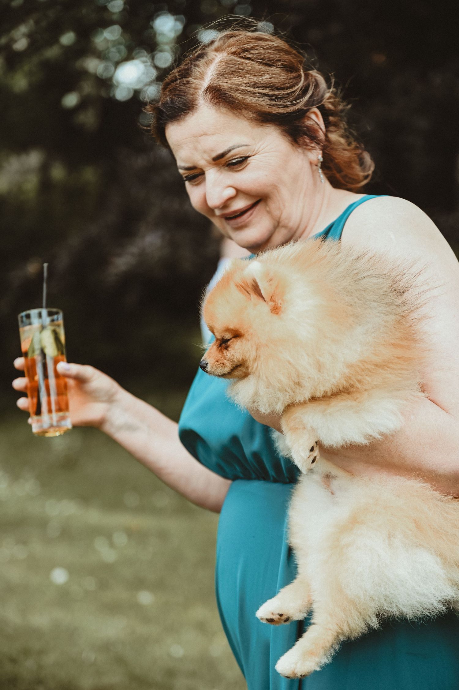 Woman in teal dress holds fluffy Pomeranian dog while enjoying a drink outdoors.