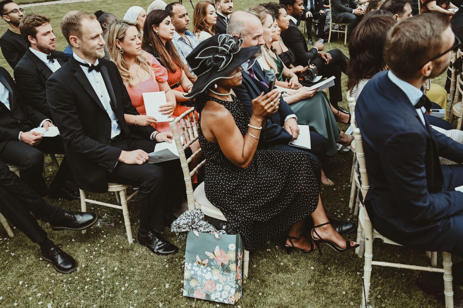 Wedding ceremony attendees seated in rows of white chairs on a manicured lawn.