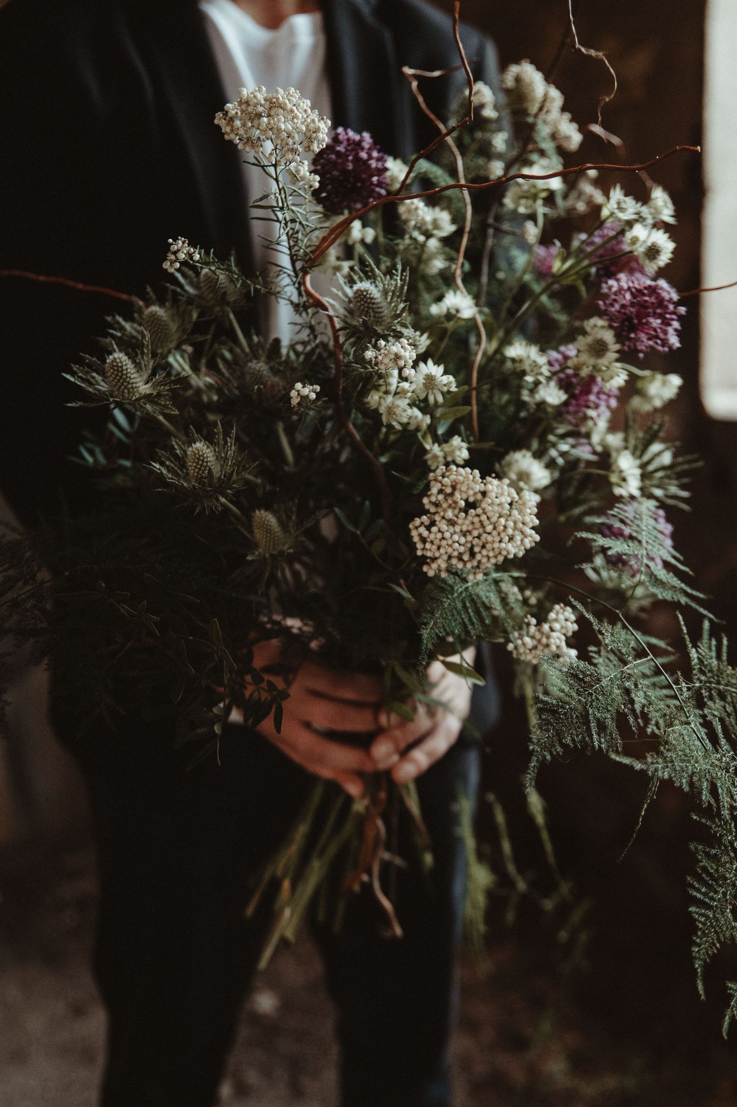 Close-up of wild botanical wedding bouquet with white flowers and greenery held against dark suit.