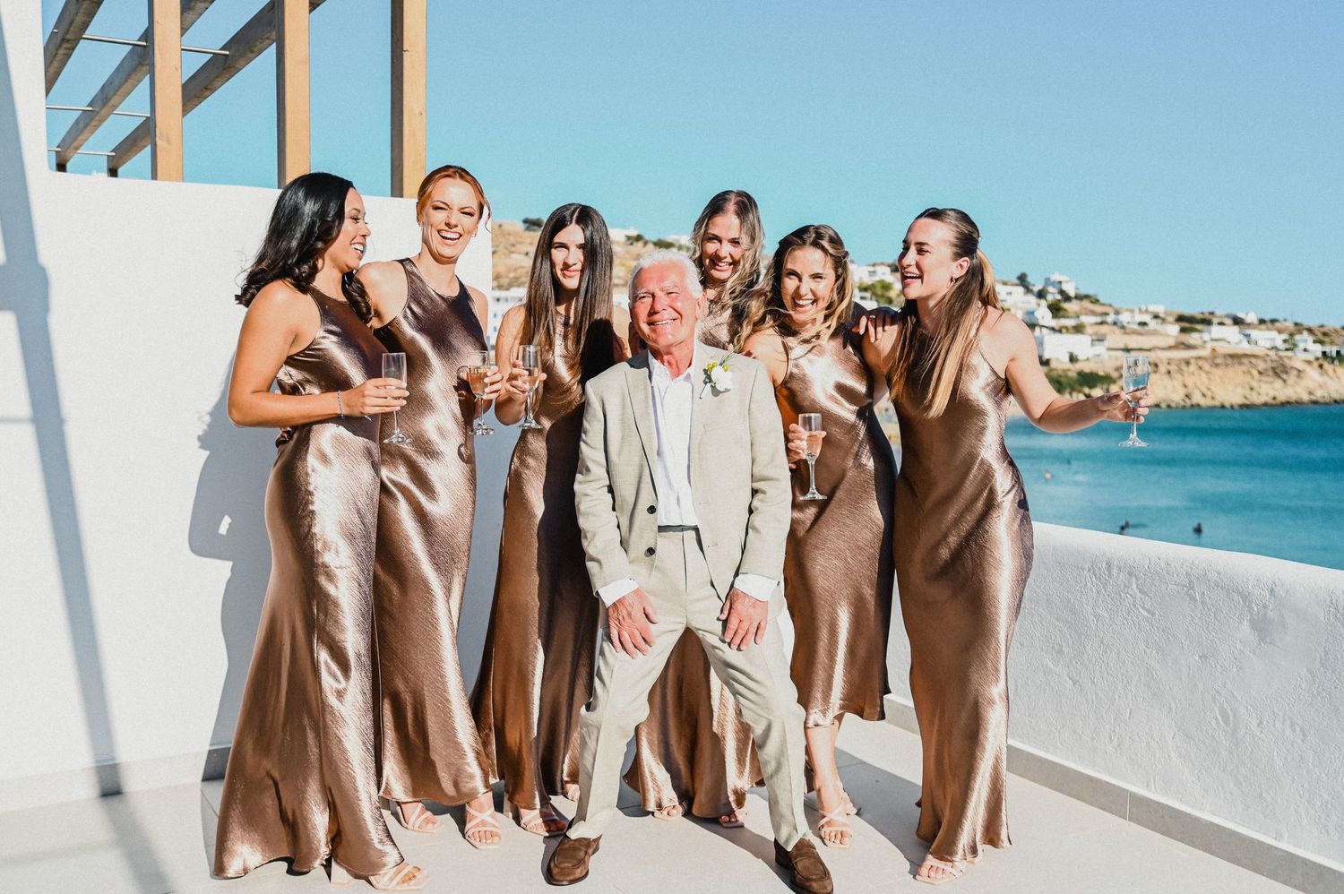 Wedding party in gold dresses poses with guest against scenic coastal backdrop on white terrace.
