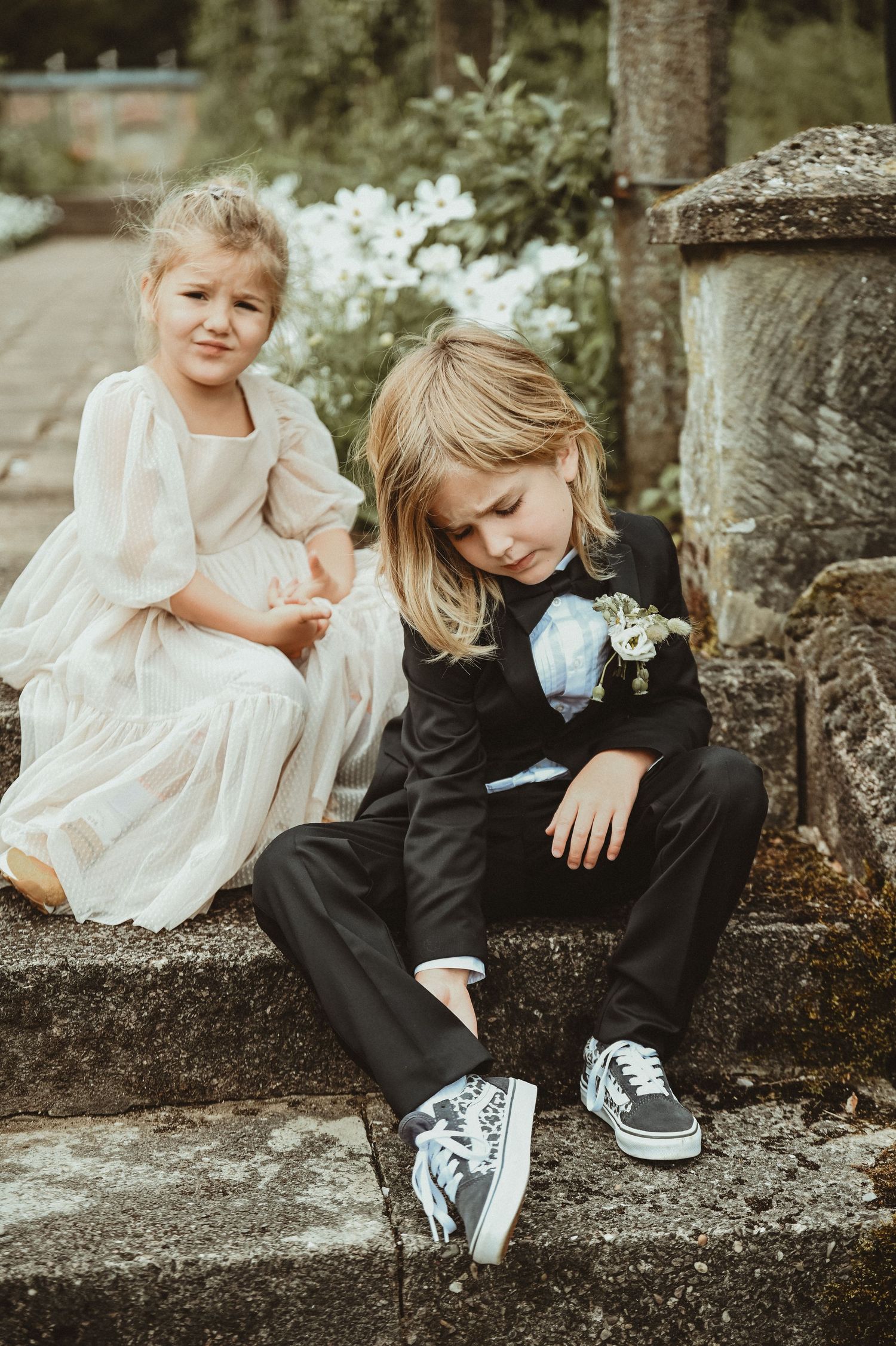 Two children in formal wear sit on stone steps during a wedding.